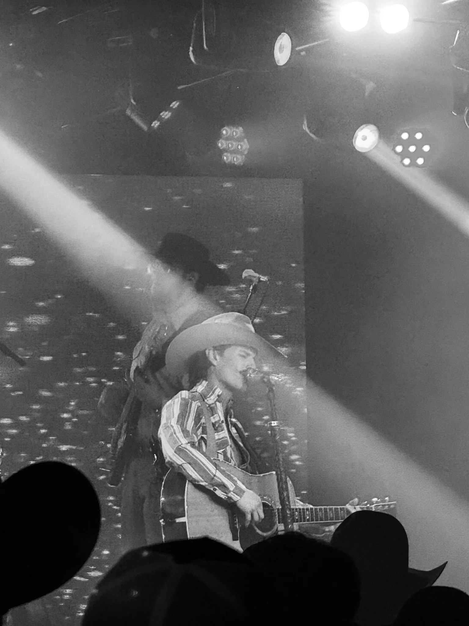 A male musician with a hat and striped shirt playing an acoustic guitar on a dimly lit stage with bright spotlights and a microphone in front of him.