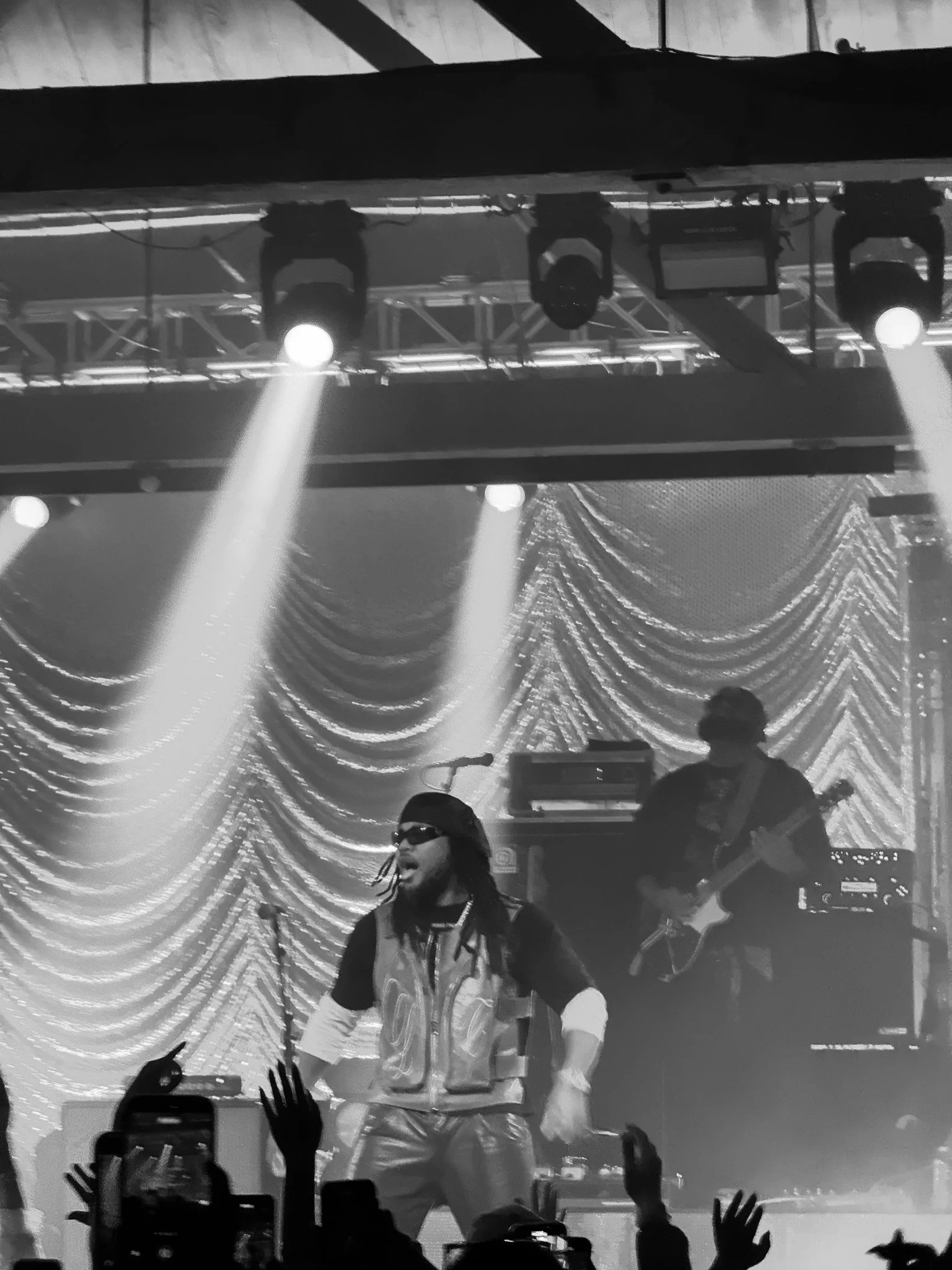 A live music concert scene in black and white featuring a singer with long hair and wearing sunglasses, engaging with the audience. The stage is illuminated with bright lights, with a backdrop of draped curtains and a metal truss structure supporting