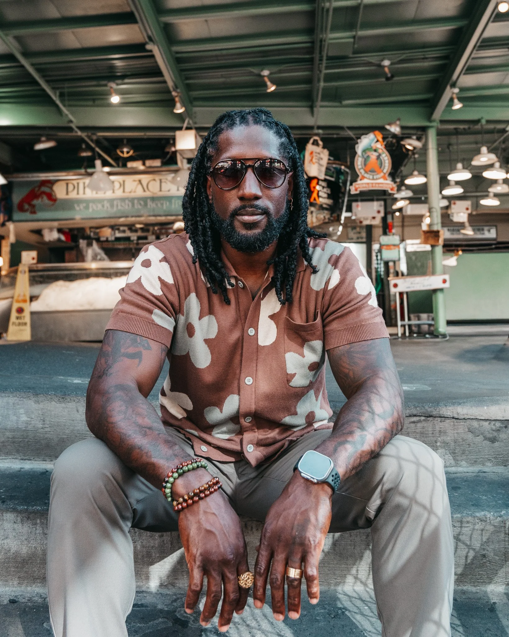 A man with braided hair, wearing sunglasses and a camouflage shirt, sitting outdoors at a table. He has multiple bracelets and rings on his hands, a smartwatch on his left wrist, and tattoos on his arms. The background shows a market or food stall wi