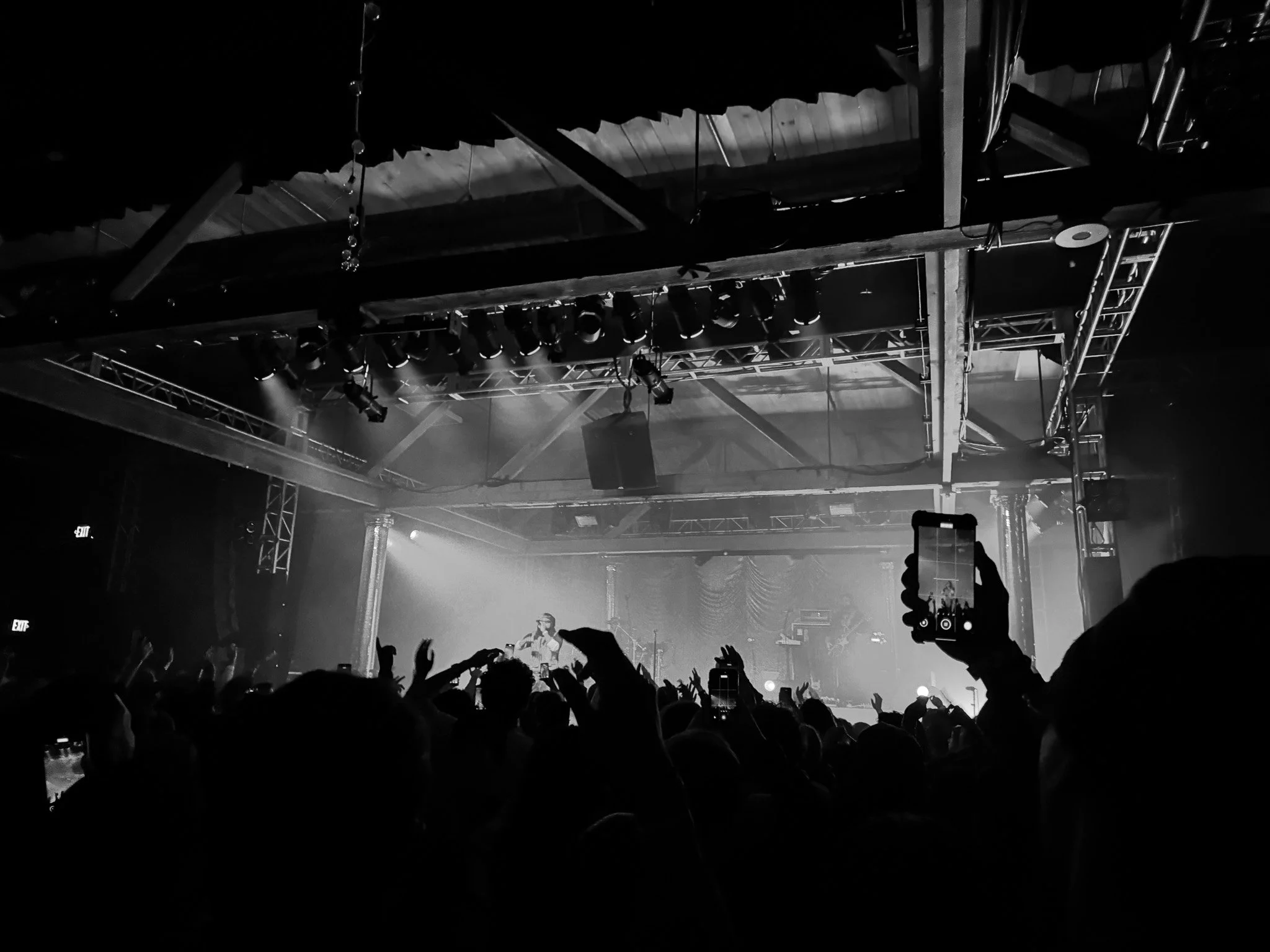 A black-and-white photo of a live concert with stage lights, a performer, and an audience raising their hands.