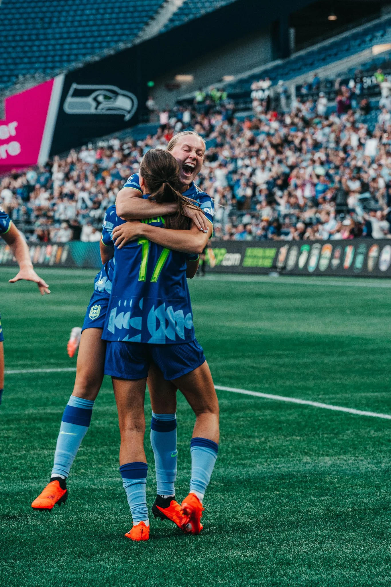 Two female soccer players in blue jerseys hugging on the field during a match, with a crowd in the stadium in the background.