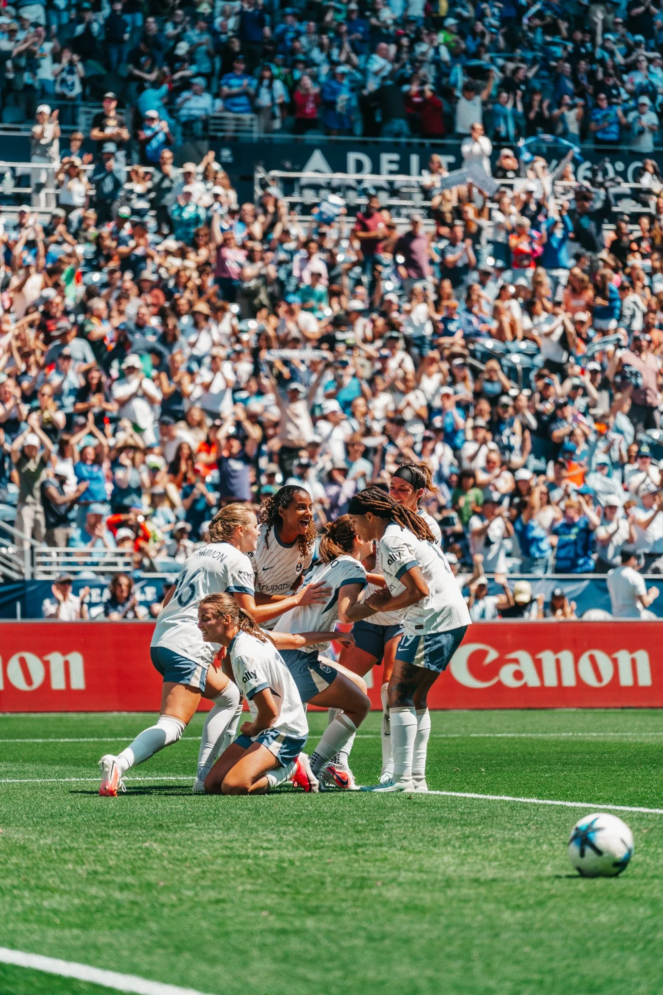 A group of female soccer players celebrating together on a green field in front of a large crowd during a match. They are wearing white jerseys and shorts, and cheering.