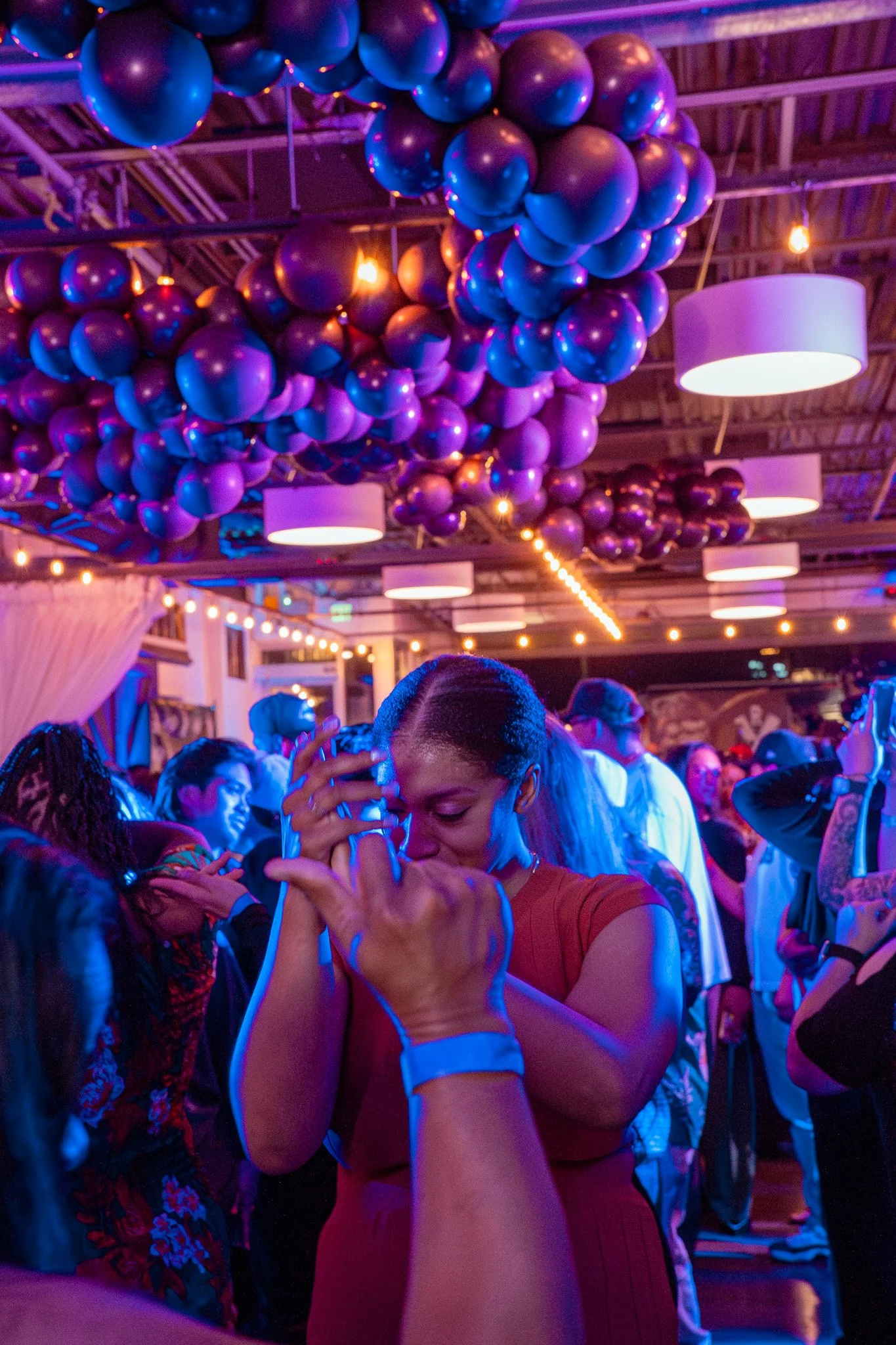 A lively party scene with couples dancing closely, illuminated by vibrant purple and blue lighting, and surrounded by festive purple balloons and string lights hanging from the ceiling.
