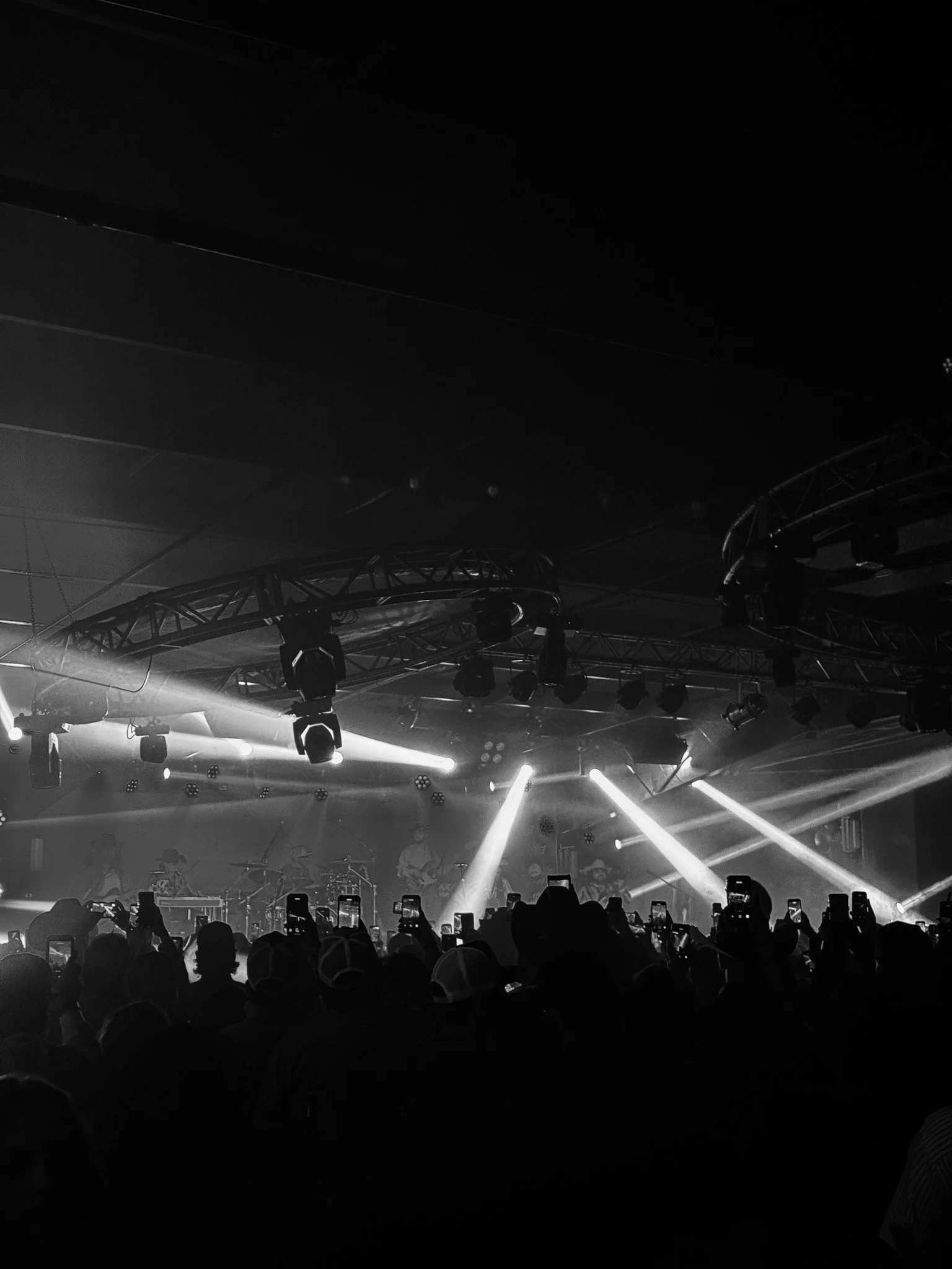 A live music concert with stage lighting and musicians performing, viewed from the audience with people taking photos.