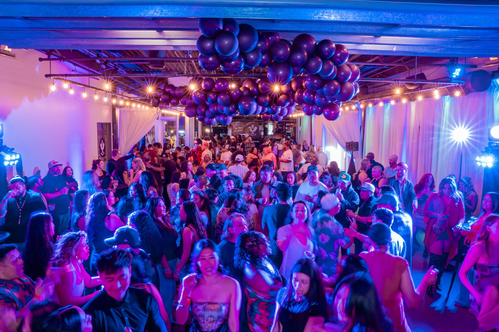 Crowd dancing at a lively indoor party with purple balloon decorations and colorful stage lighting.