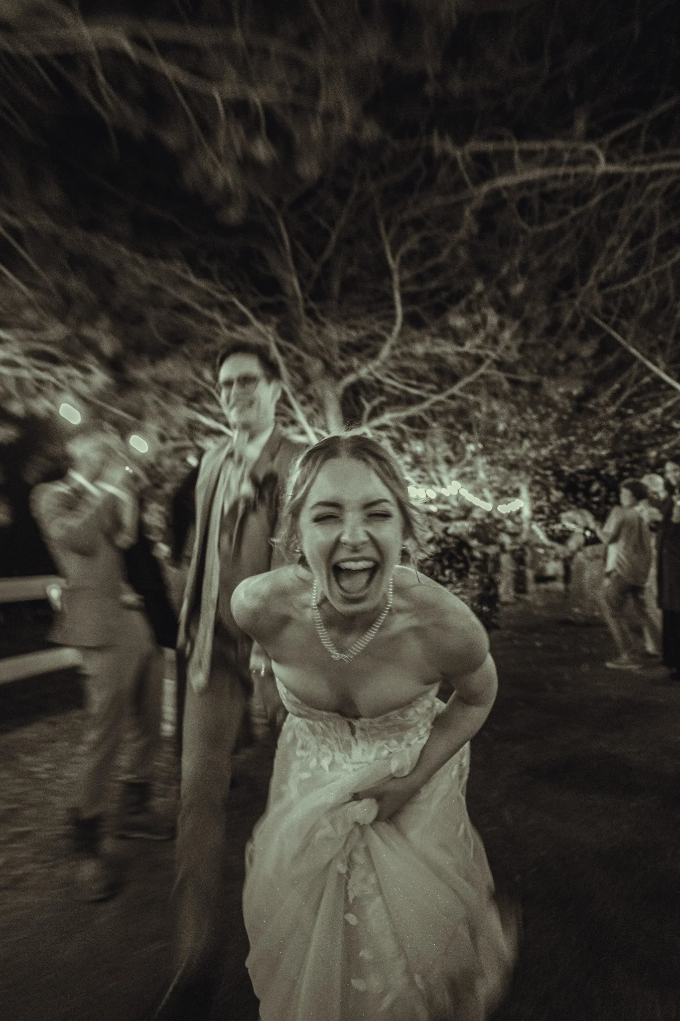 A woman in a wedding dress laughing and bending forward, surrounded by people at an outdoor event with a large tree in the background.