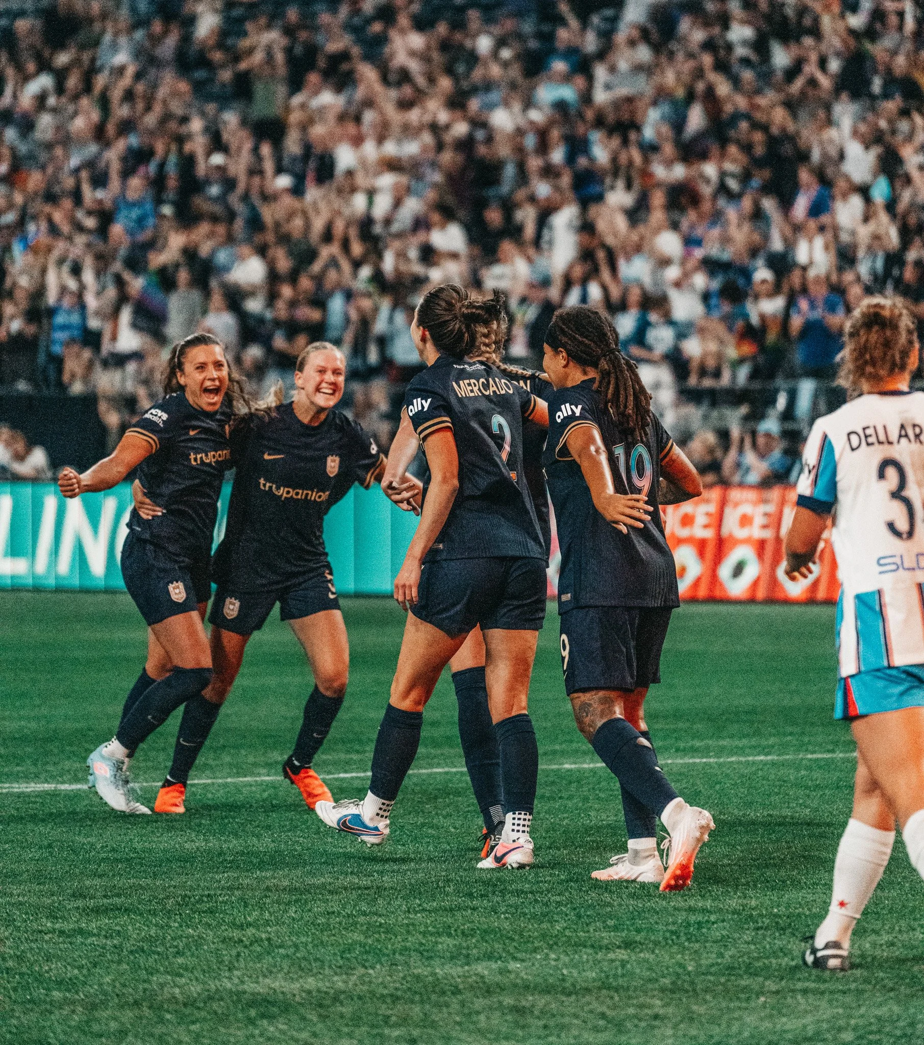A group of female soccer players celebrating on the field while a large crowd watches in the stands.