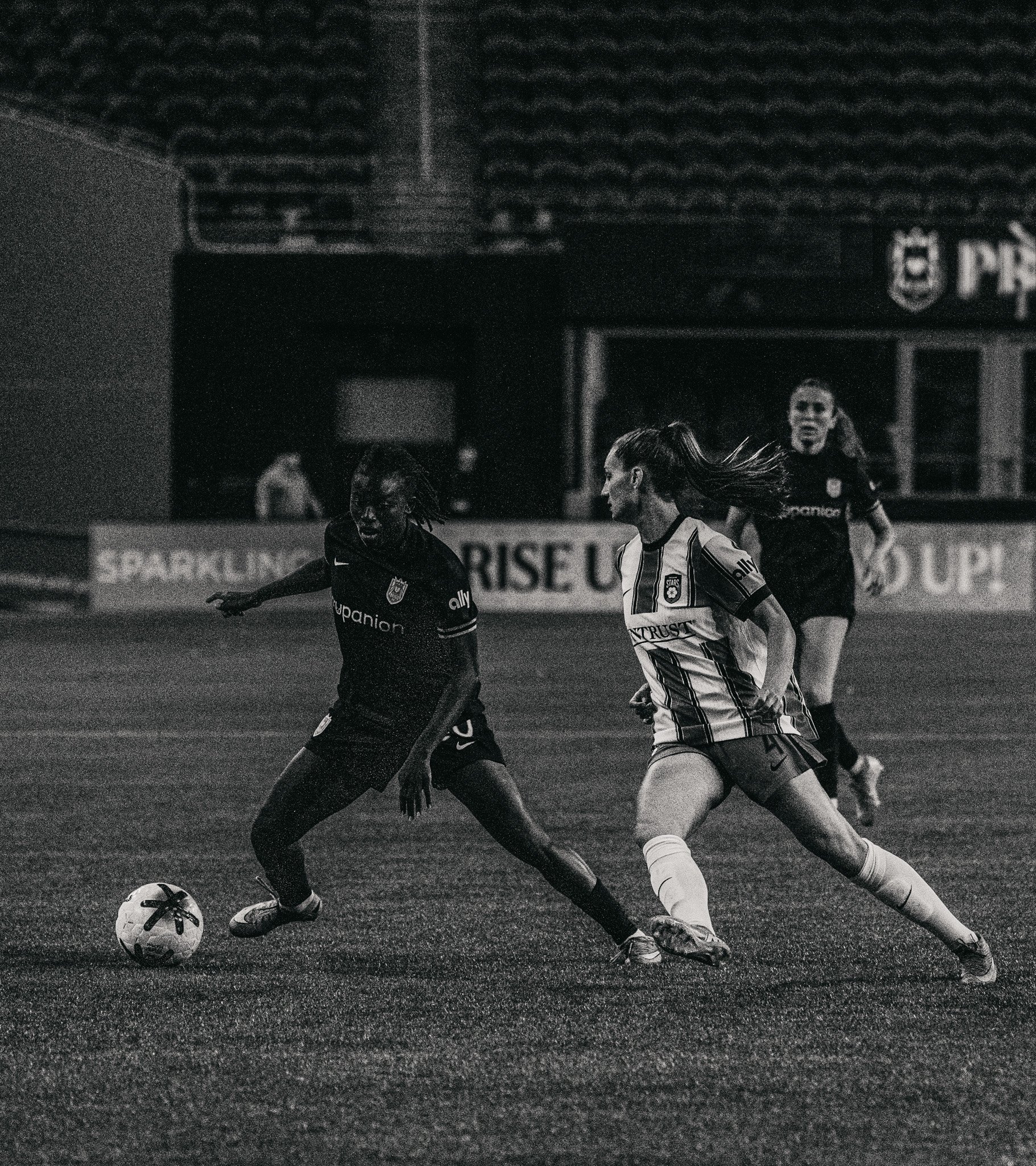 Two female soccer players compete for the ball during a game in a stadium. One player in a dark jersey is kicking the ball, and the other in a white striped jersey is running alongside. A referee and other players are visible in the background.