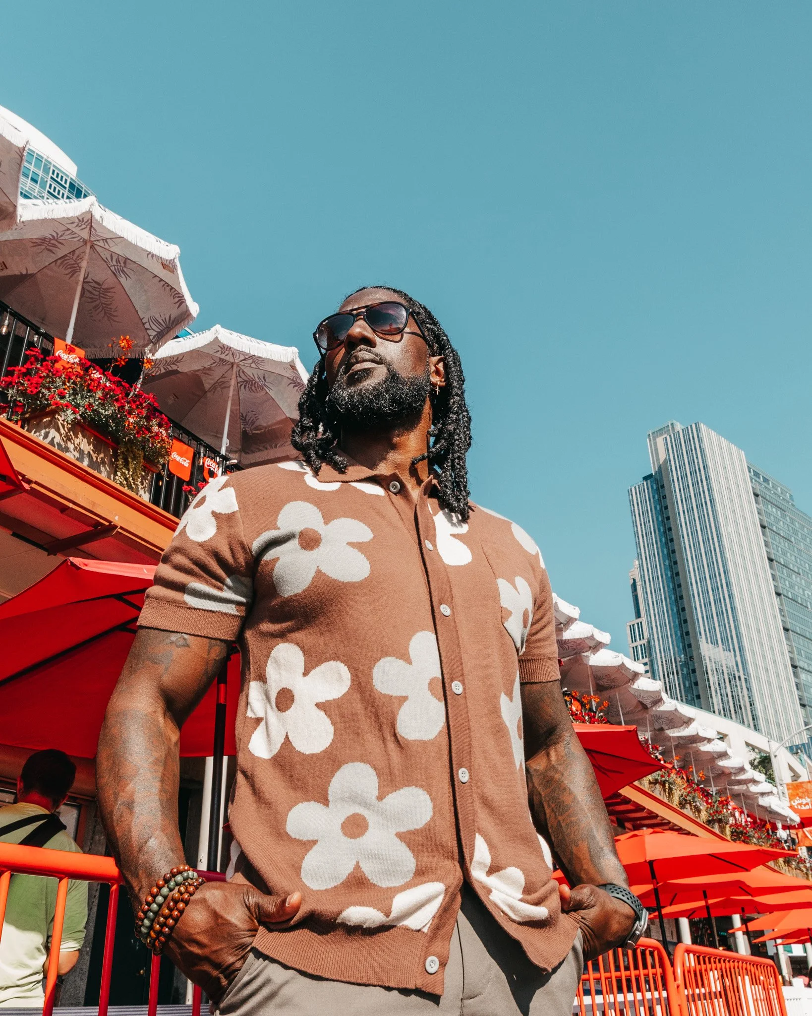 A man with long dreadlocks and sunglasses standing outdoors amidst orange umbrellas, modern tall buildings, and a clear blue sky.