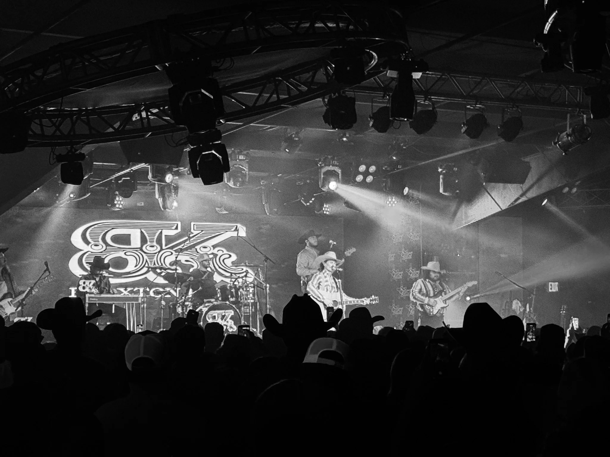 Black and white photo of a crowded concert stage with band members performing. Stage is filled with lighting and musical equipment, audience silhouettes visible in the foreground.