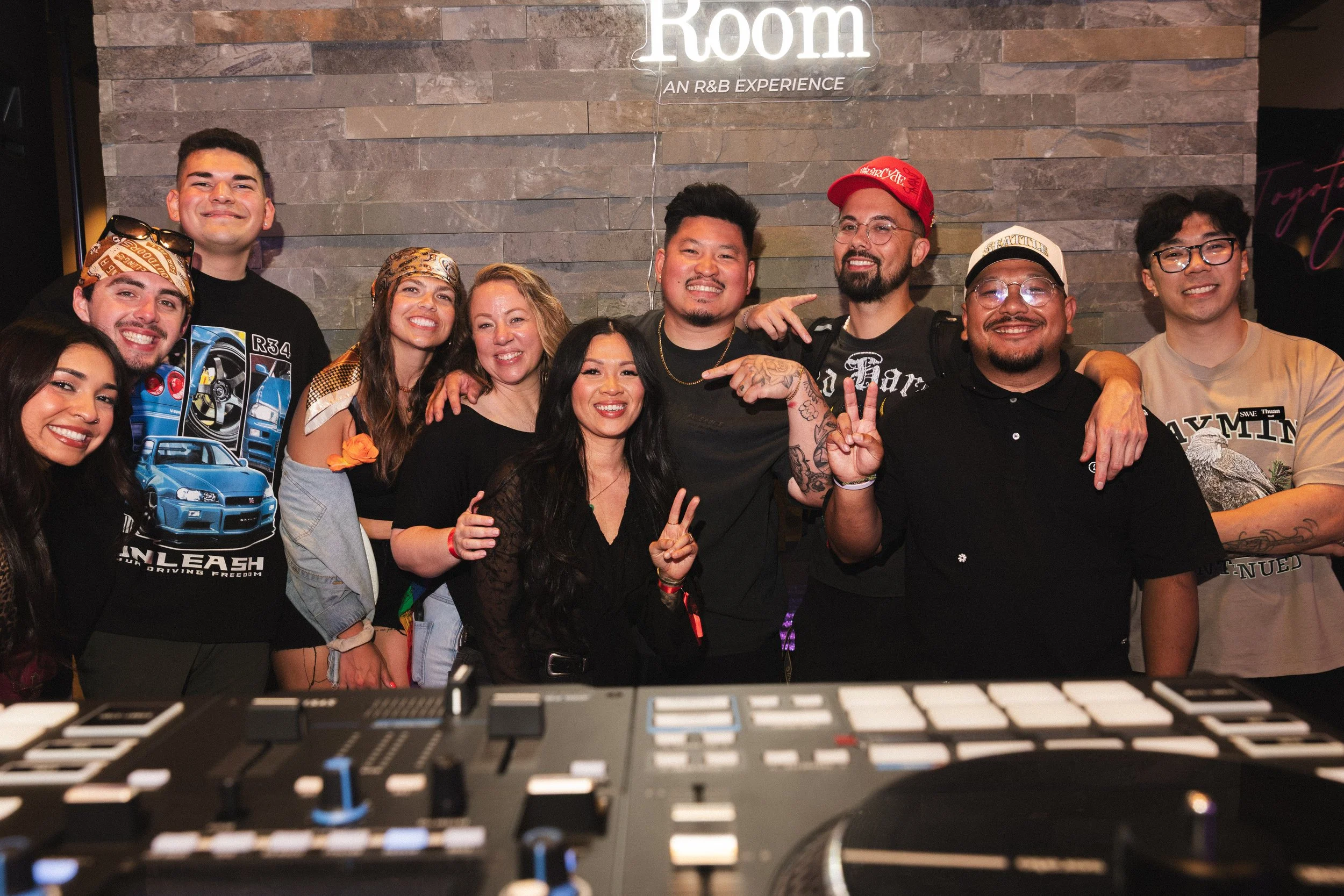 A group of diverse smiling people posing together in front of a brick wall with a neon sign that reads 'Room: An R&B Experience' in a dimly lit setting, with a keyboard or DJ equipment in the foreground.