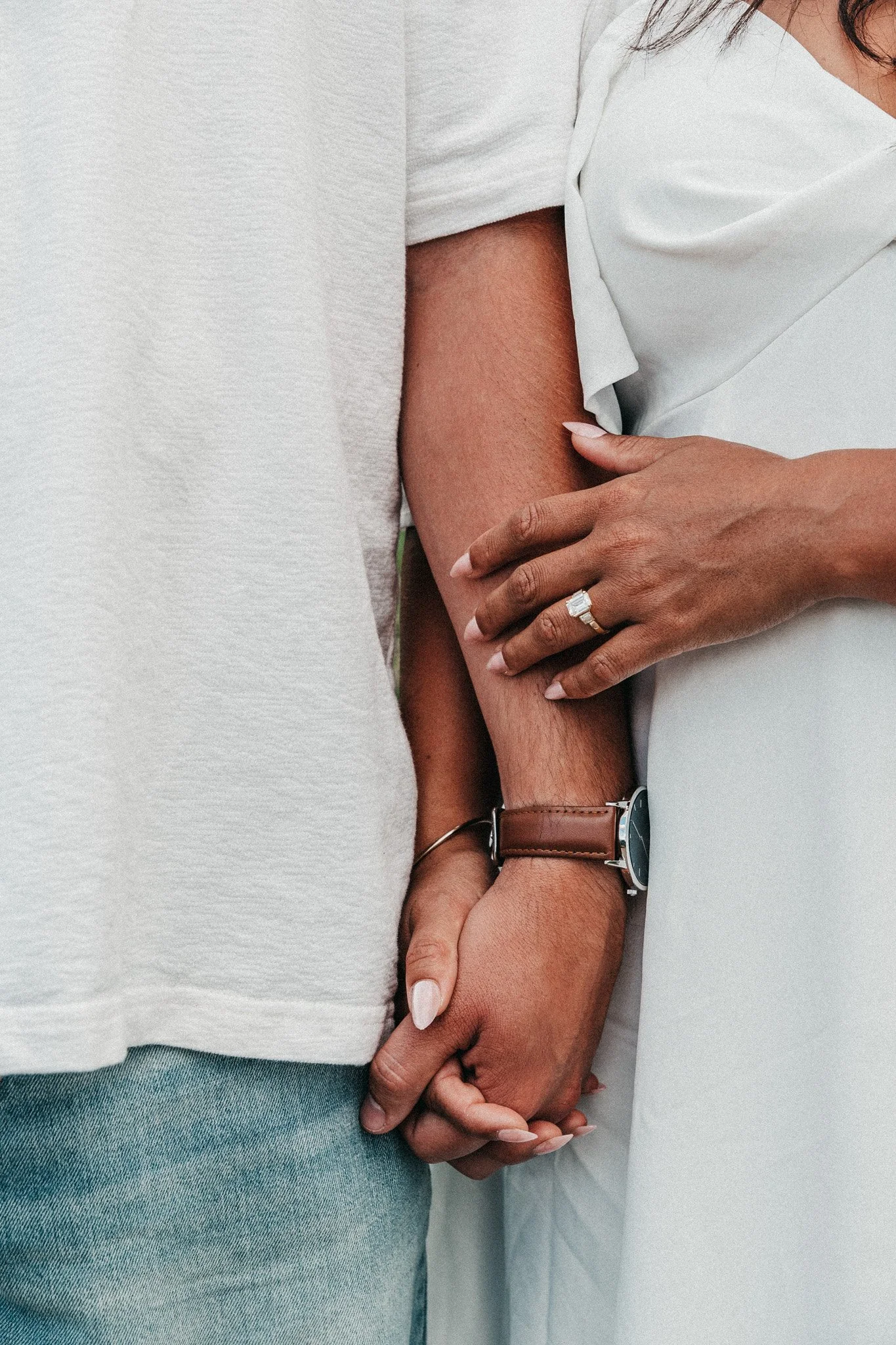 Two individuals holding hands, one wearing a white top and the other a gray shirt, with close-up focus on their hands and arms.