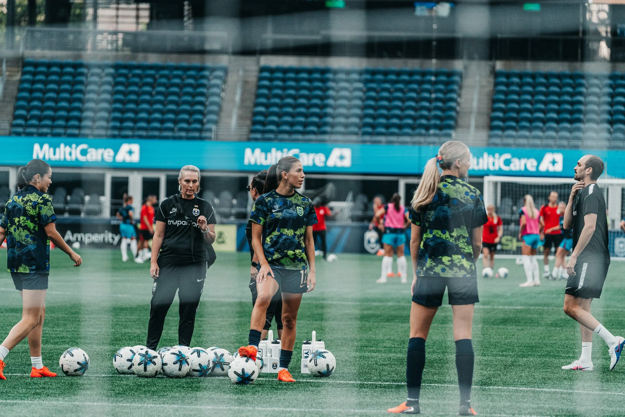Women soccer players on the field during practice, some are standing while others are walking near a line of soccer balls and water bottles.