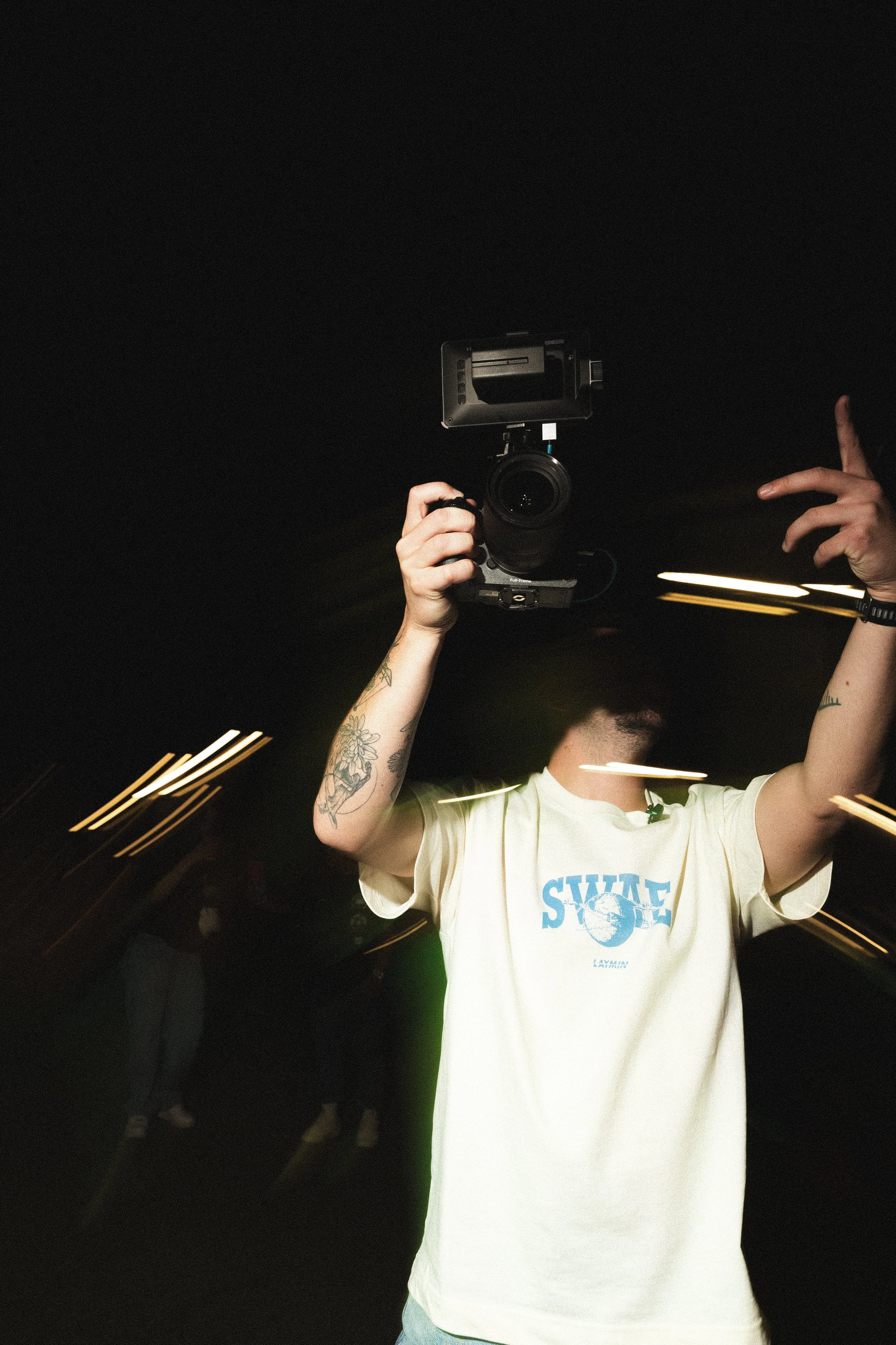 A person wearing a cream-colored T-shirt with the word 'SWAG' and a globe graphic, holding a camera with both hands, and pointing with their right hand, taken in a dark environment with blurred lights and other people in the background.