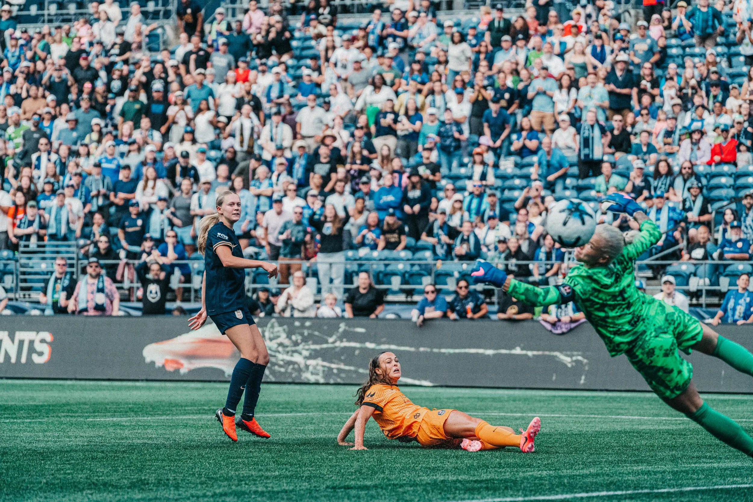 A soccer match with players and spectators, showing a goalkeeper in green diving to make a save, a player in orange on the ground, and another player in blue nearby
