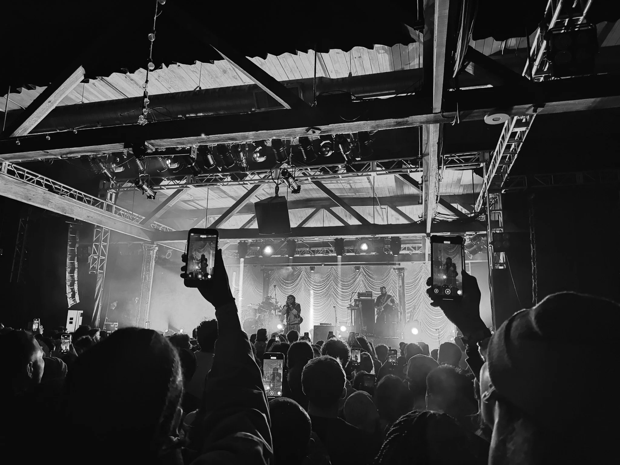 Black and white photo of a live concert with stage lighting, drum set, and musicians in the background. Audience members are visible, some holding phones to record the performance.