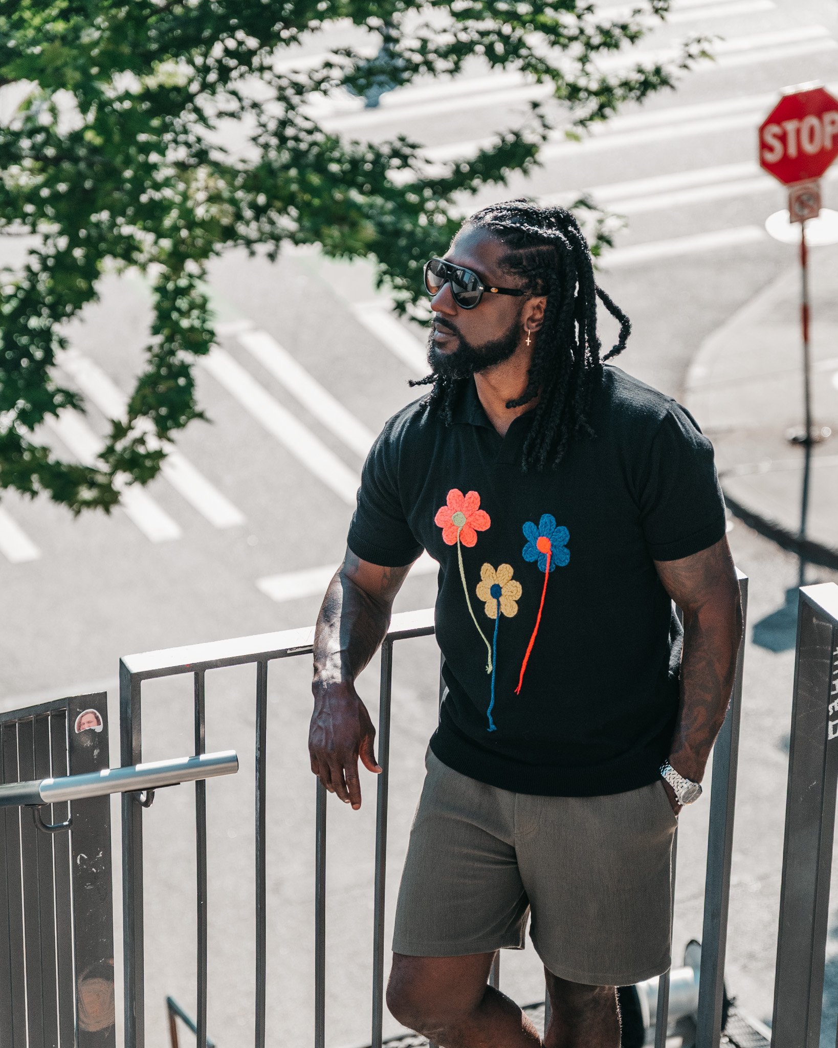 A man with braided hair, sunglasses, and earrings standing outdoors near a metal barricade. He is wearing a black shirt with colorful flower designs and gray shorts. There is a stop sign in the background and a tree with green leaves.