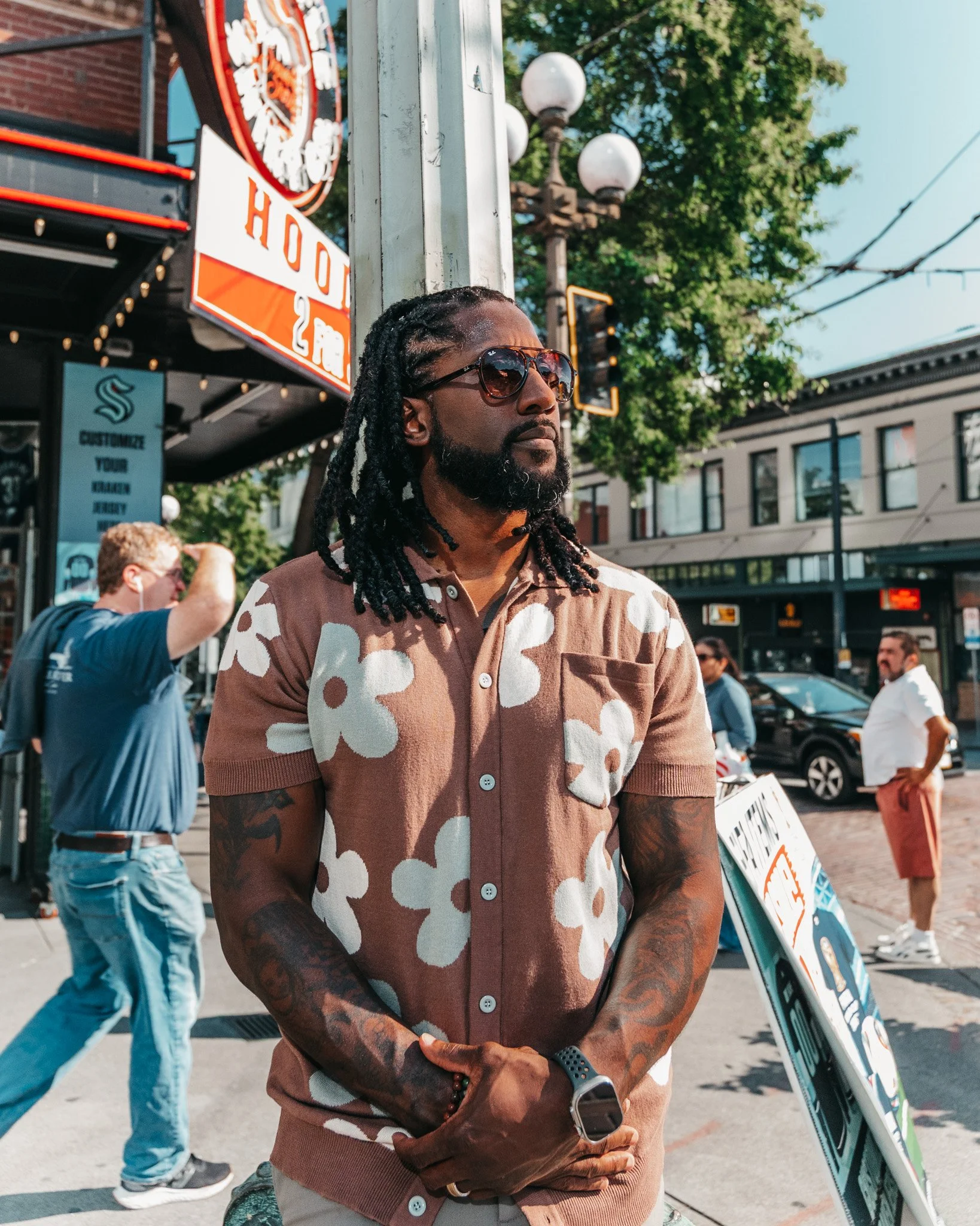 A man with long dreadlocks, wearing sunglasses, a beige and brown patterned shirt, and a smart watch, stands outdoors in an urban setting with trees, buildings, and a group of people in the background. There are signs and street lights nearby.
