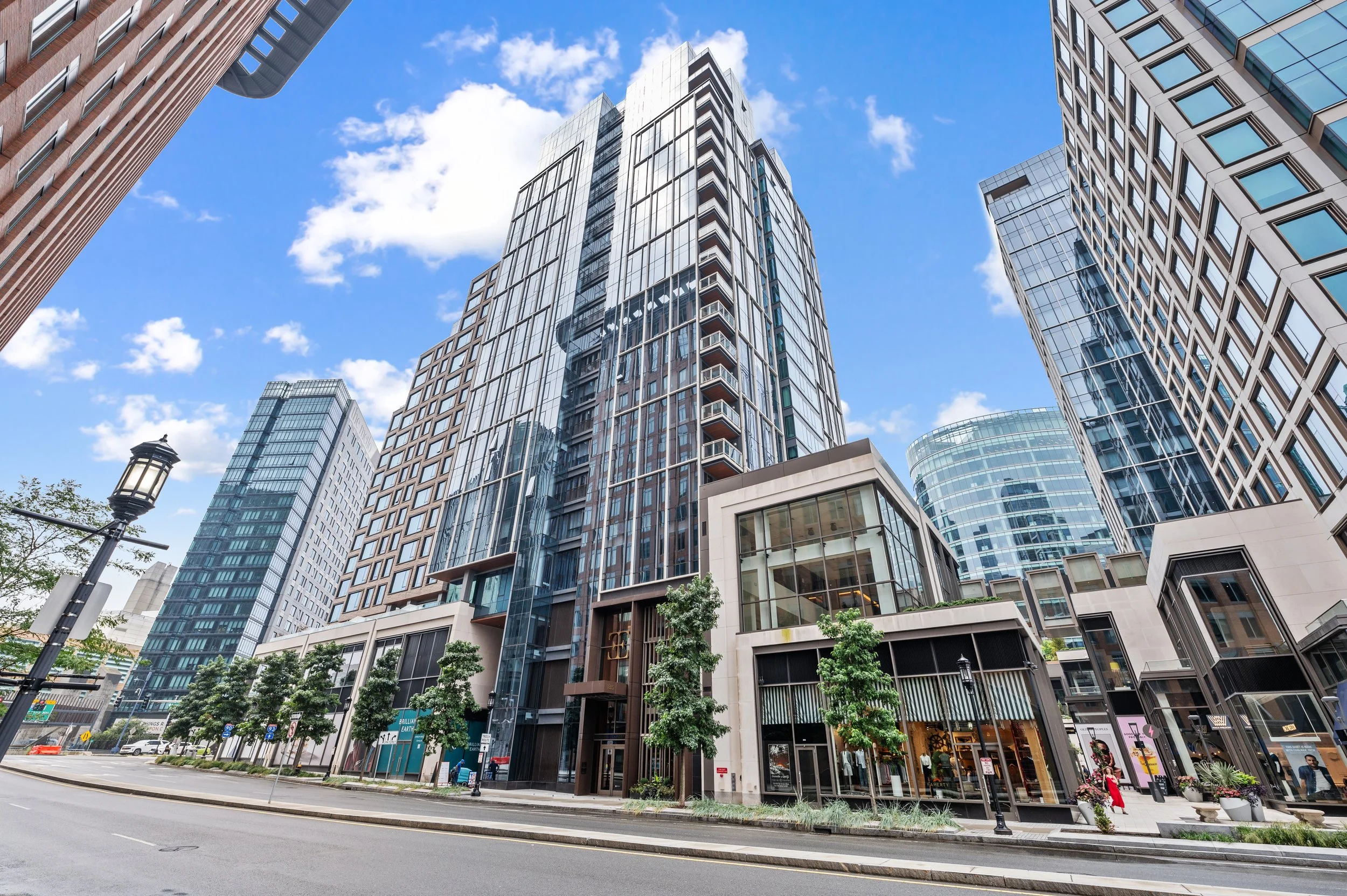 Tall modern glass building in the Seaport District in Boston lining a city street with trees, a lamppost, and pedestrians under a partly cloudy blue sky.