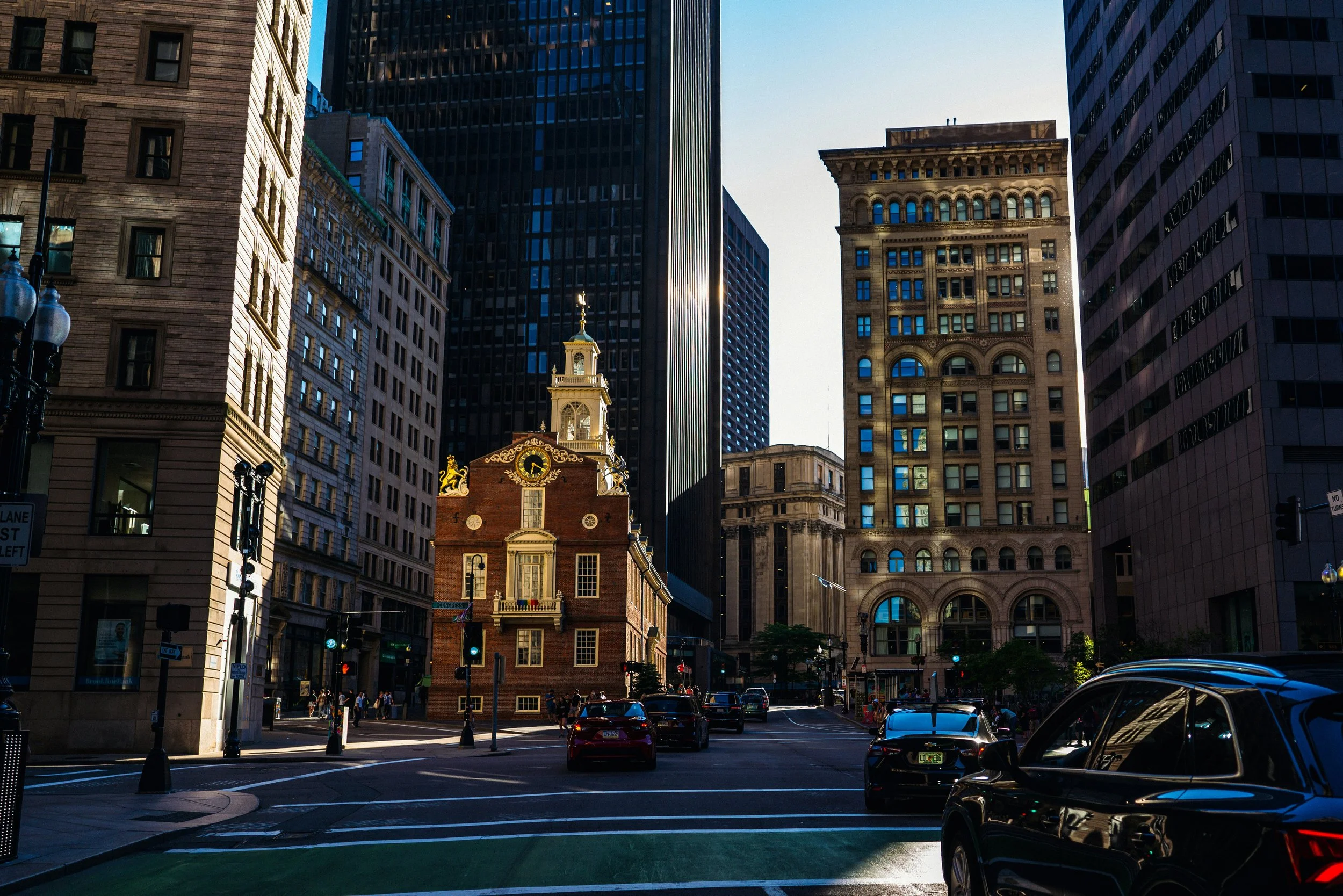 Boston's city street scene with tall modern buildings and an historic building with a clock tower in the center, cars parked and walking pedestrians.