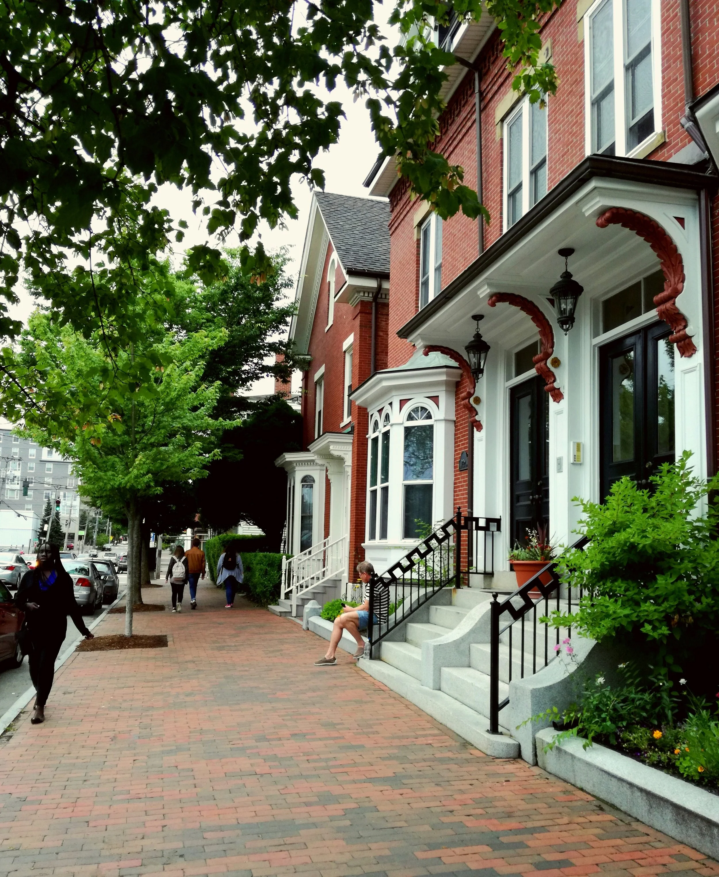 A row of red-brick townhouses with black doors and decorative trim, set along a tree-lined sidewalk in Boston's Charlestown neighborhood.