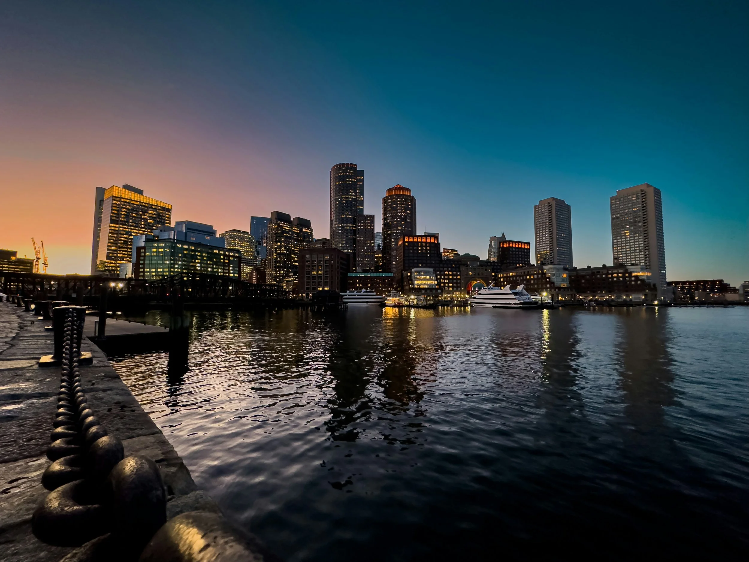 Boston city skyline at dusk with tall buildings reflecting in the water, boats docked at the harbor, and a sky transitioning from orange to deep blue.