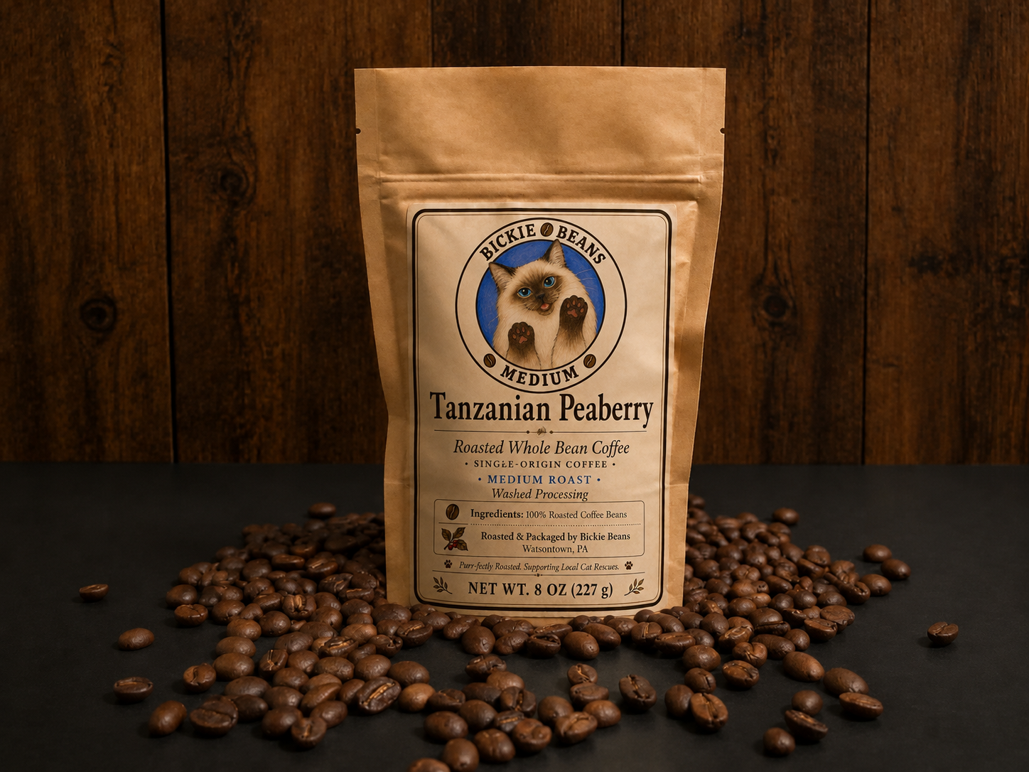 A beige coffee bag labeled "Tanzanian Peaberry" with a cat illustration, surrounded by roasted coffee beans on a dark surface against a wooden background.