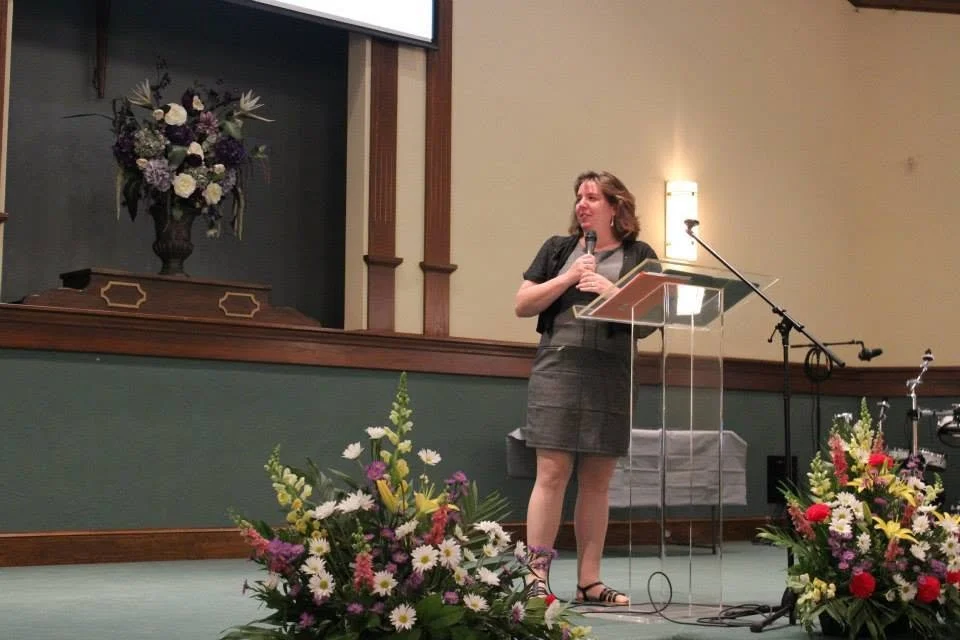 A woman standing at a clear podium with a microphone, giving a speech or presentation in a decorated indoor venue with floral arrangements on the stage.