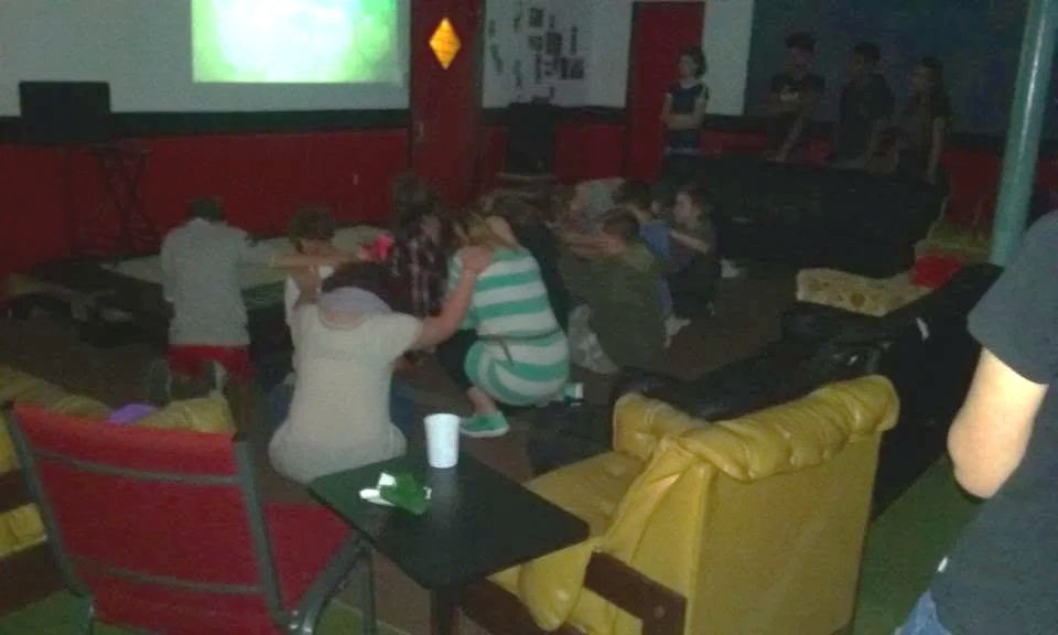A group of children and teenagers in a dark room, possibly a gaming or entertainment room, sitting on sofas and chairs, watching a screen, with some standing and gathered around.