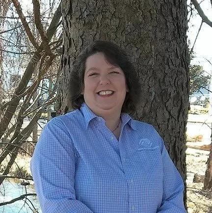 Woman smiling outdoors in front of a large tree, wearing a blue checkered shirt.