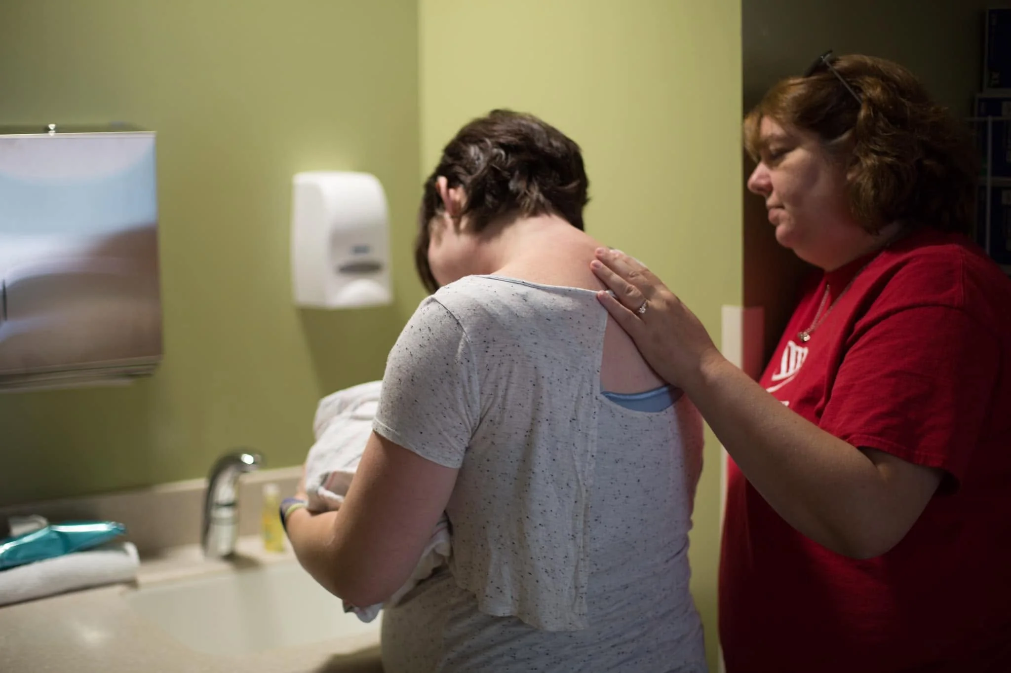 A woman in a red shirt gently caresses a patient's shoulder in a hospital room.