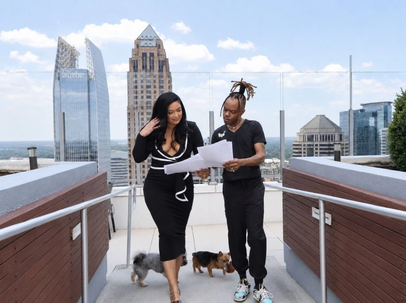 Two people, a woman in a black dress and a man with dreadlocks in black sneakers and shirt, are looking at papers on a rooftop with a city skyline in the background. Two dogs are walking beside them.