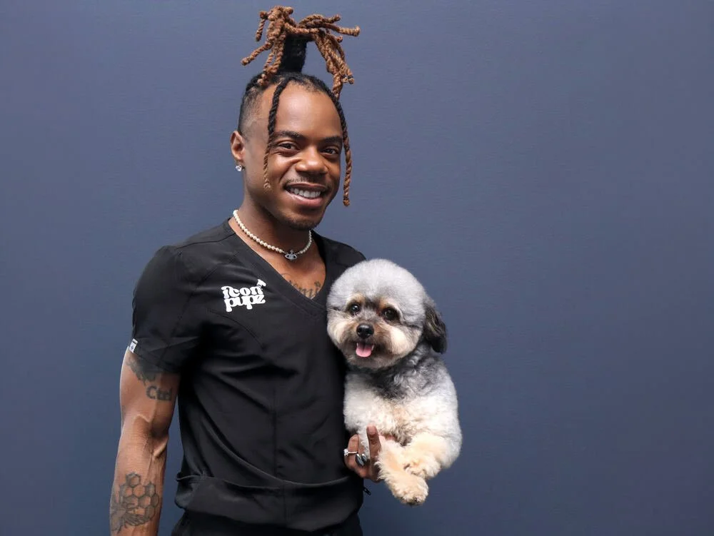 A man with styled dreadlocks, wearing a black shirt and jewelry, holding a small gray and white dog, standing against a plain dark blue background.