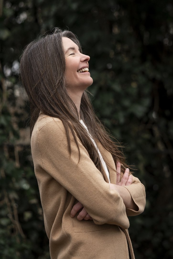 A woman with long brown hair smiling with eyes closed, arms crossed, wearing a tan coat outdoors.