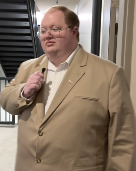 James Risner with glasses and red hair, wearing a beige suit and a white shirt, standing indoors and holding a microphone.