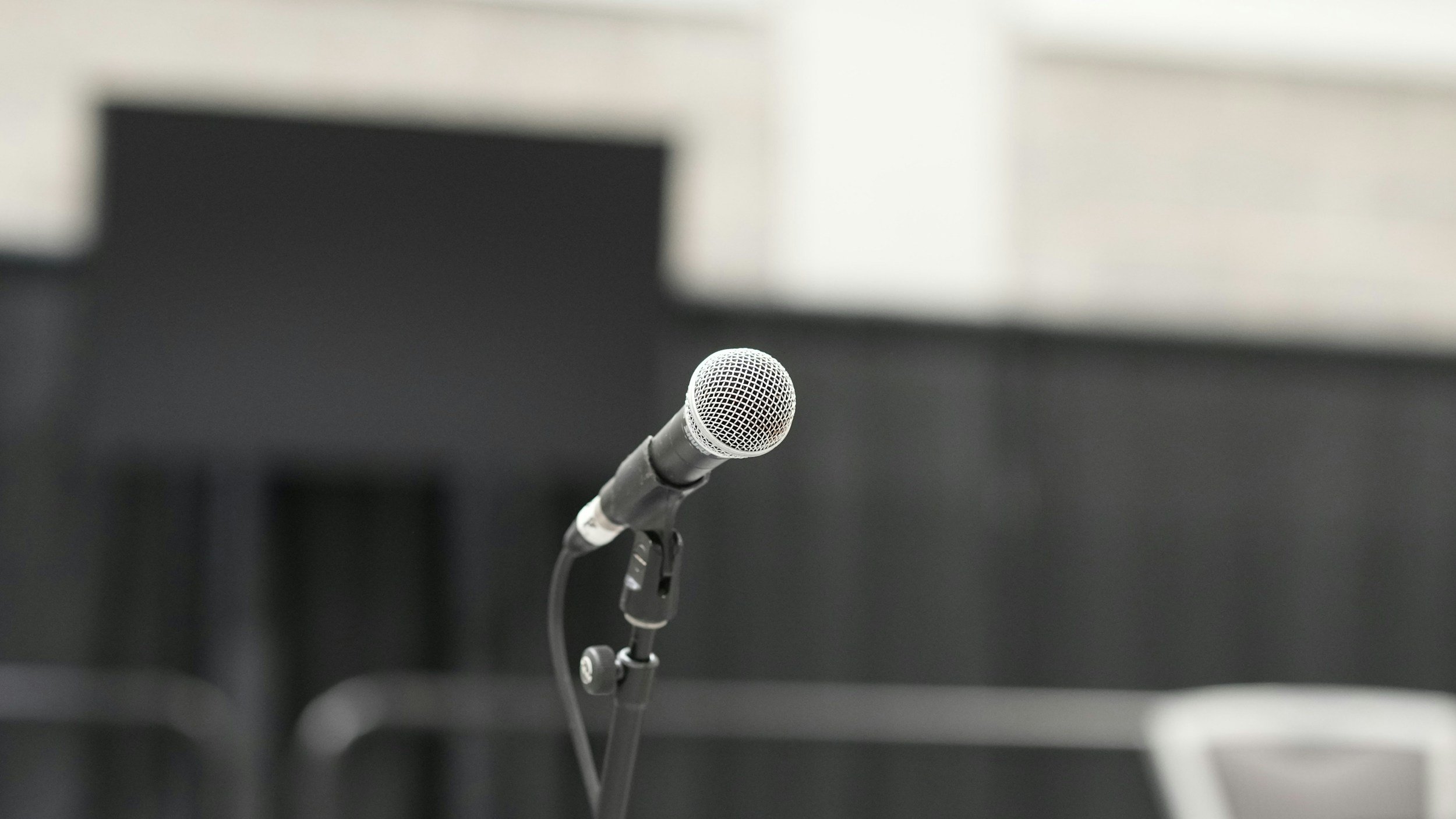A close-up of a silver microphone on a stand with a blurry office building as the background.