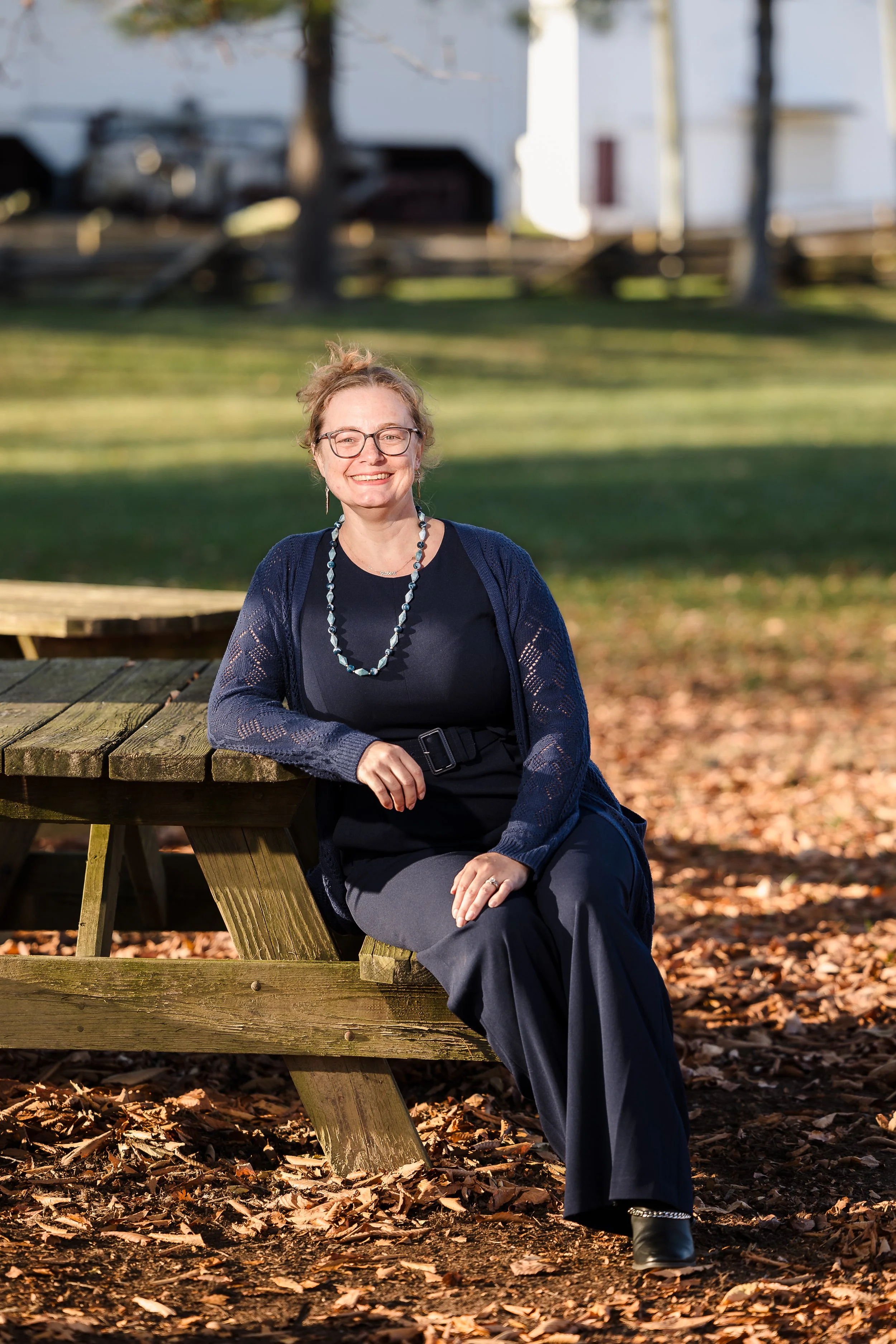 A woman with curly hair and glasses sitting on a wooden picnic table outdoors, smiling at the camera, with trees and autumn leaves on the ground in the background.