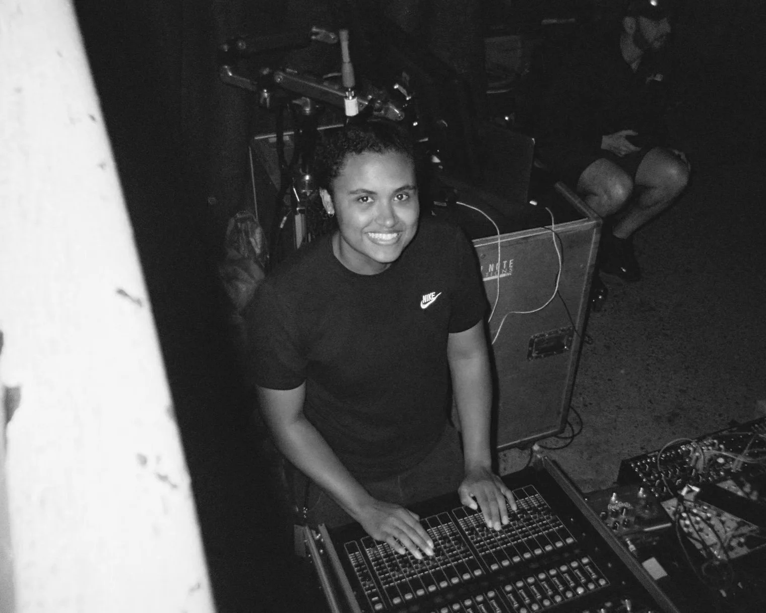 A woman with curly hair, smiling, wearing a black Nike t-shirt, operates a mixing console in a dimly lit setting with DJ equipment around her.