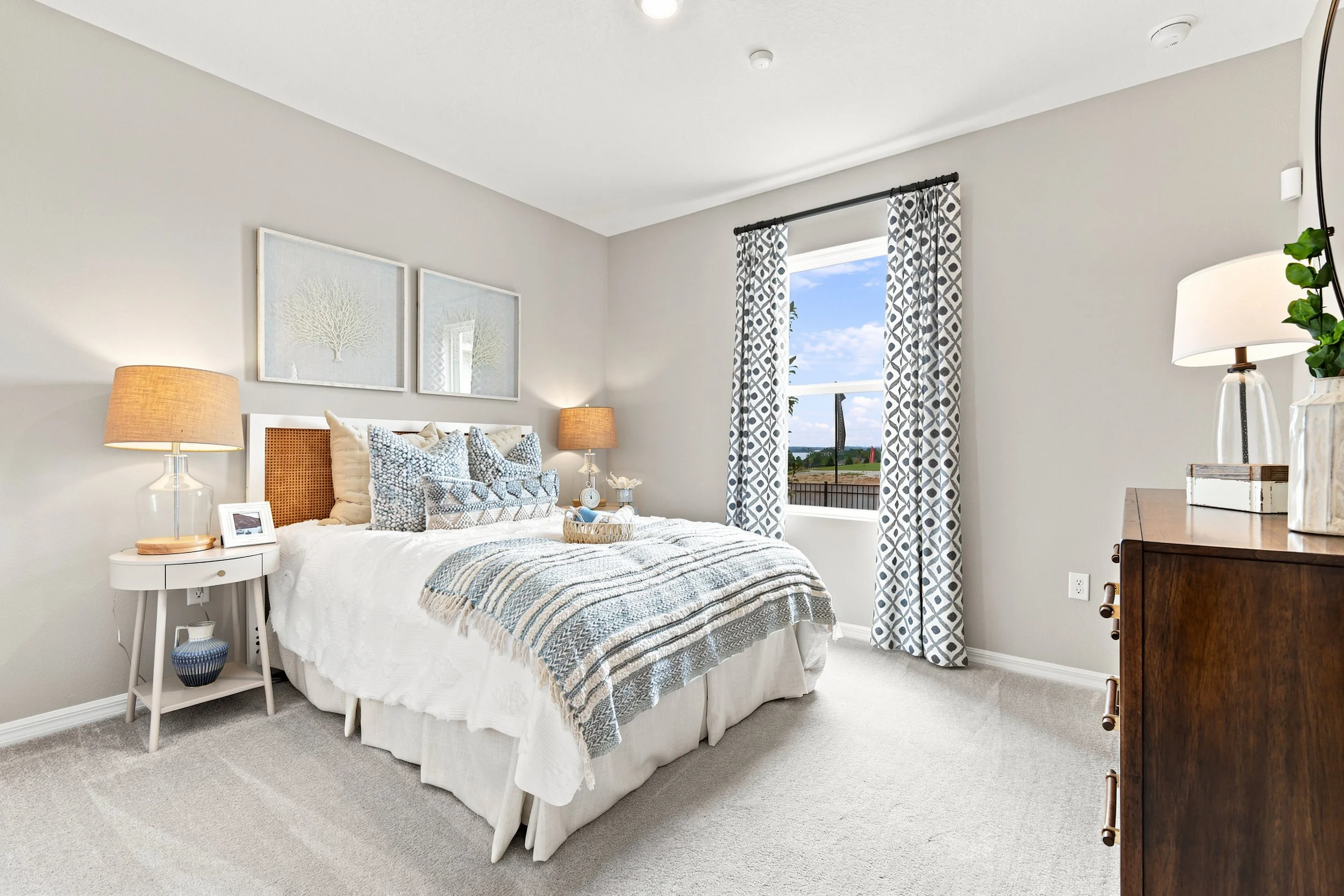 A bedroom with a white bed, two matching bedside lamps, and a window with patterned curtains showing a view of a partly cloudy sky and landscape.