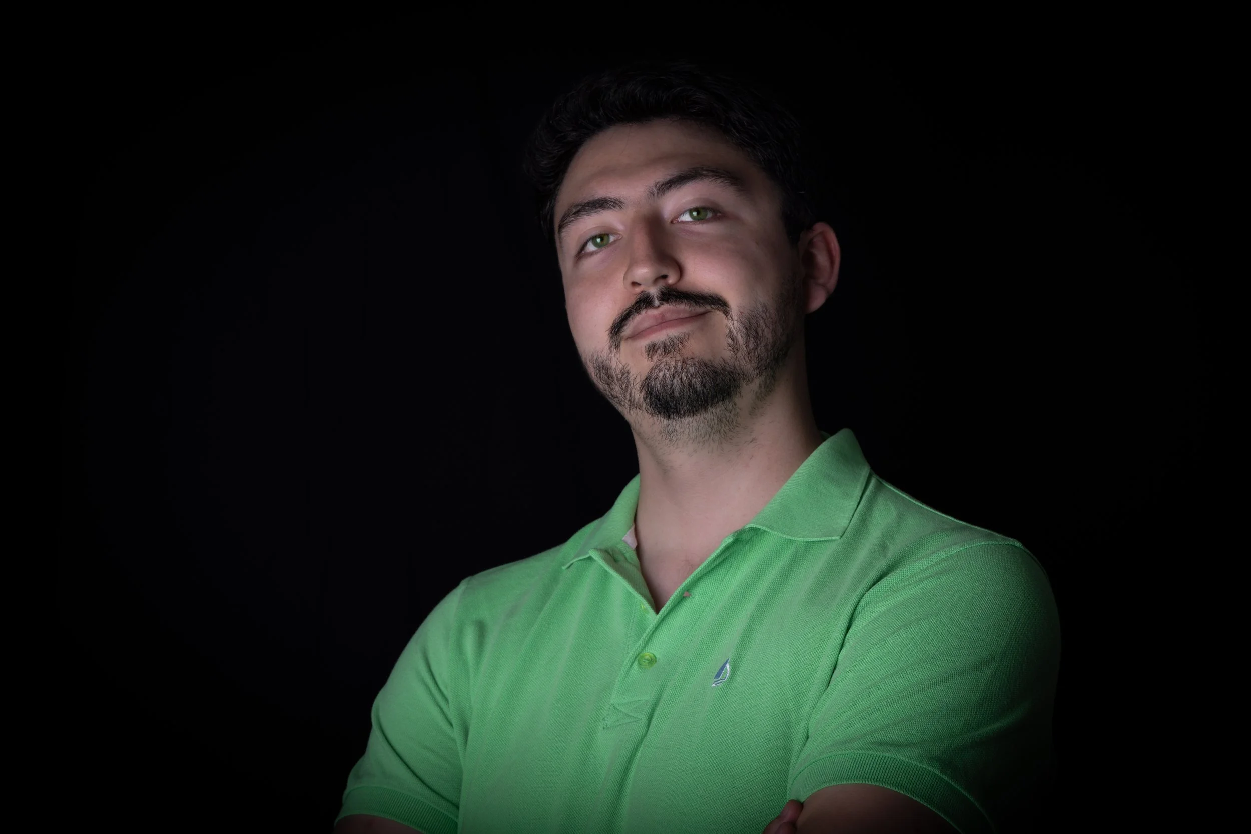 A young man with dark hair, a beard, wearing a bright green polo shirt, smiling slightly against a black background.