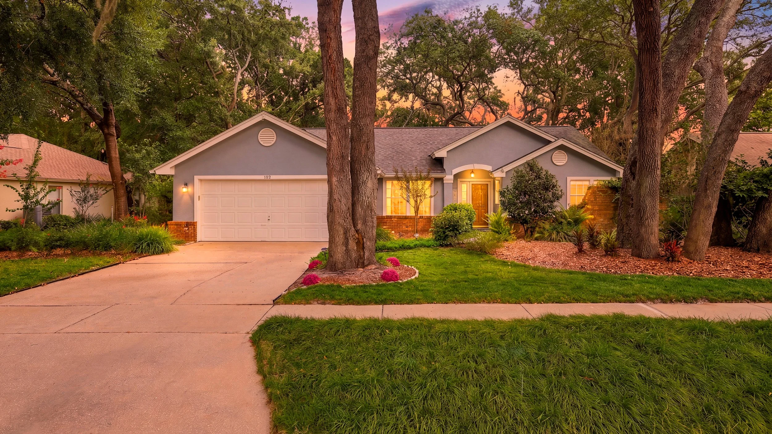 A modern house with a light blue exterior, a white garage door, and a wooden front door, surrounded by trees and landscaped garden during sunset.