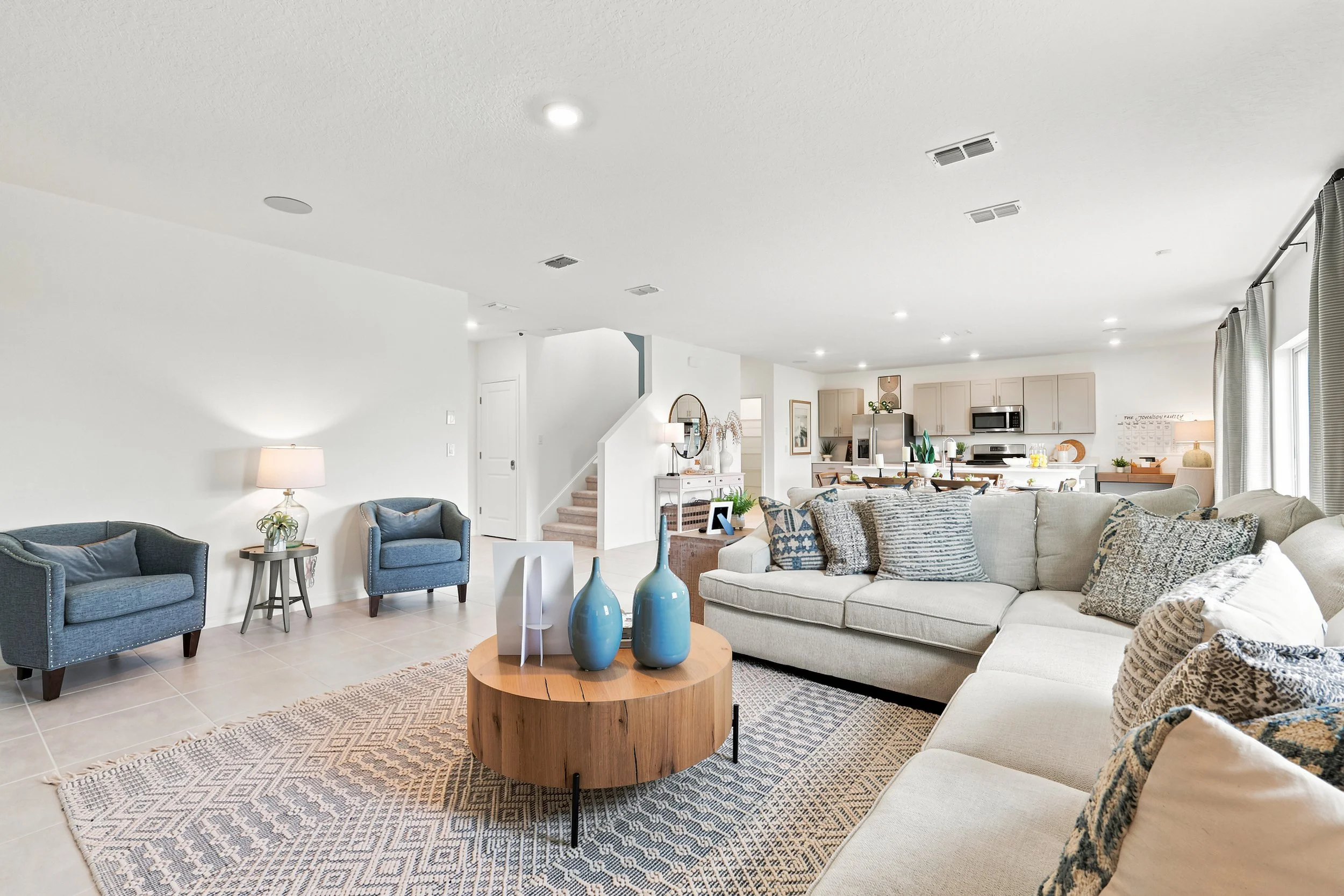 Bright, spacious living room with white walls, beige tiled floor, beige sectional sofa with patterned pillows, blue accent chairs, round wooden coffee table with three blue vases, and a patterned rug. Open-concept kitchen in the background with light wood cabinetry and stainless steel appliances. Large windows with light curtains.