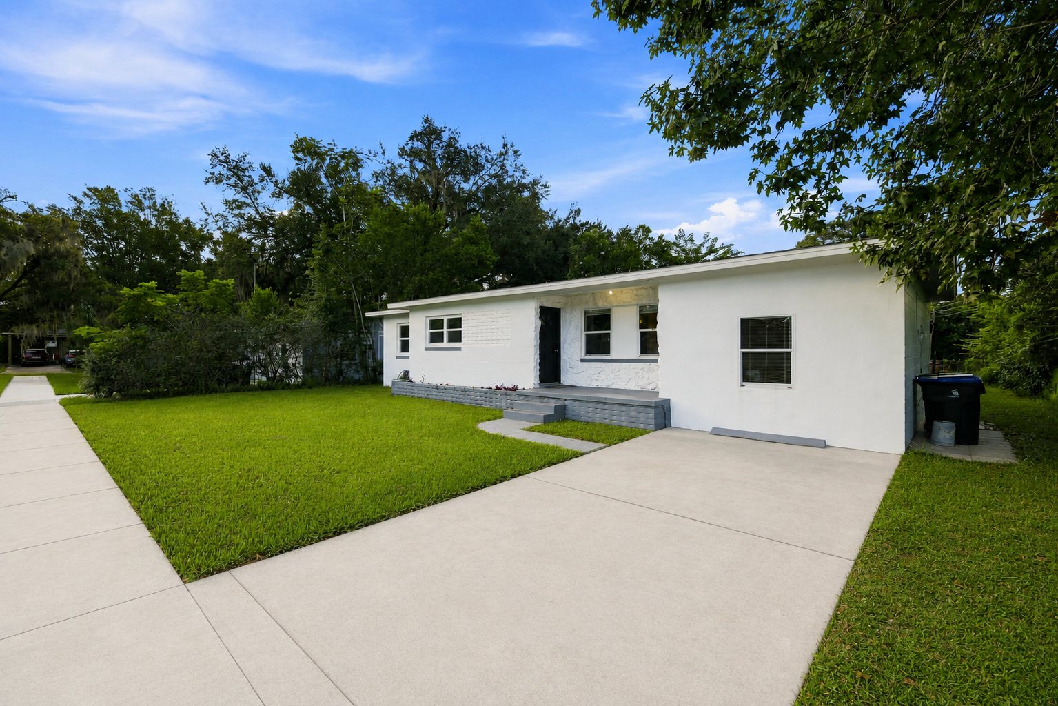 Front view of a white house with a small porch, green lawn, and concrete driveway under a partly cloudy sky.