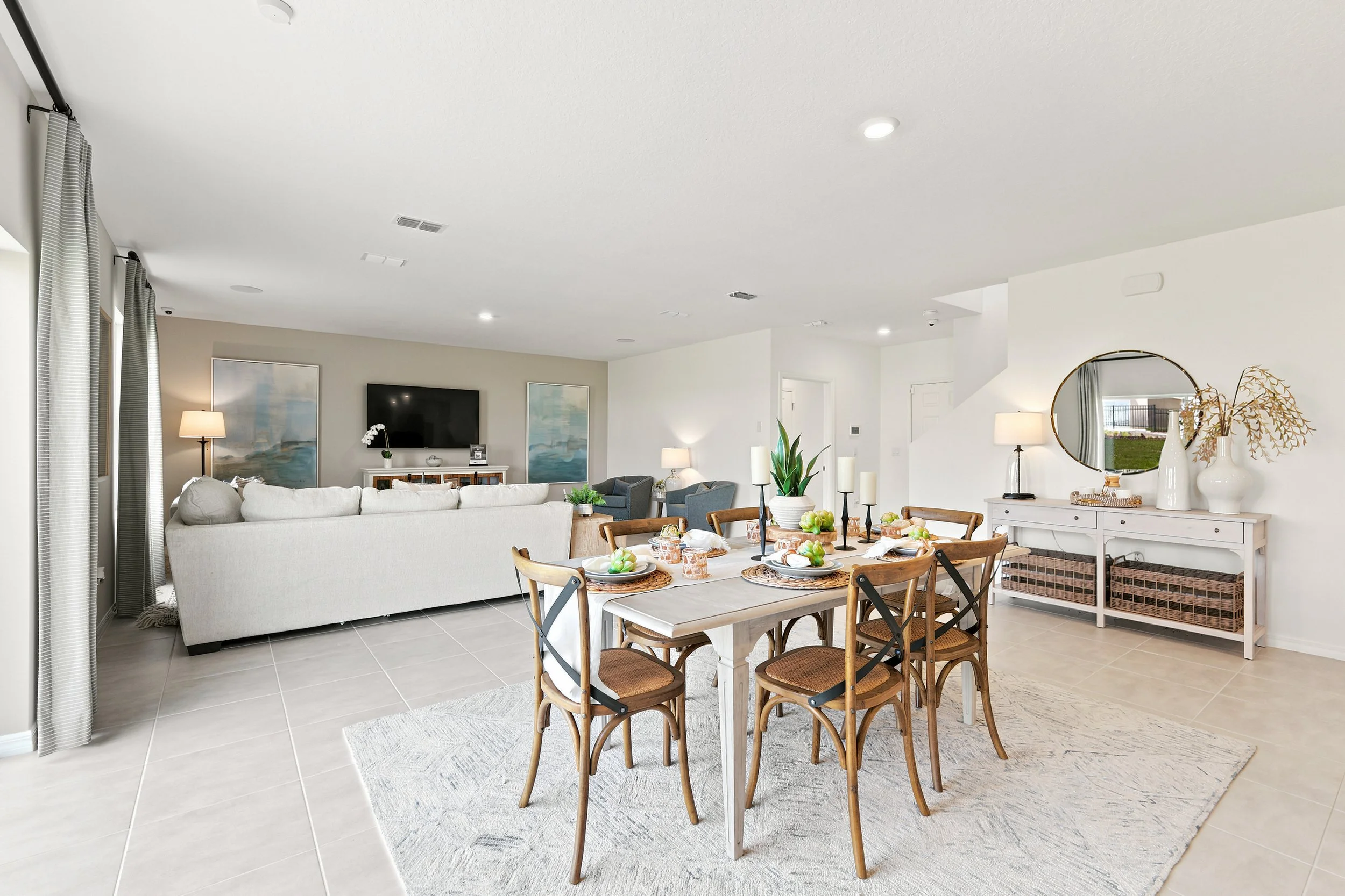 Open-concept living and dining room with white walls, grey tiled flooring, a white sofa, a dining table with wooden chairs, and decorative items like vases and artwork.