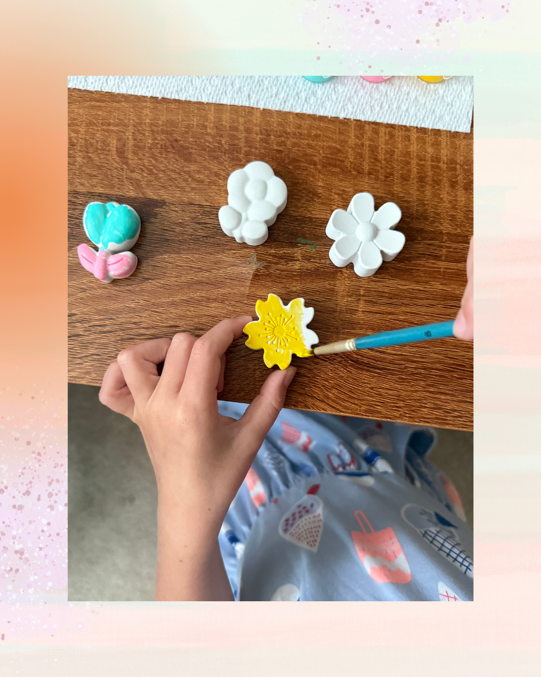 Child painting a yellow flower on a ceramic flower decoration, with three other unpainted and semi-painted ceramic flowers on a wooden surface.