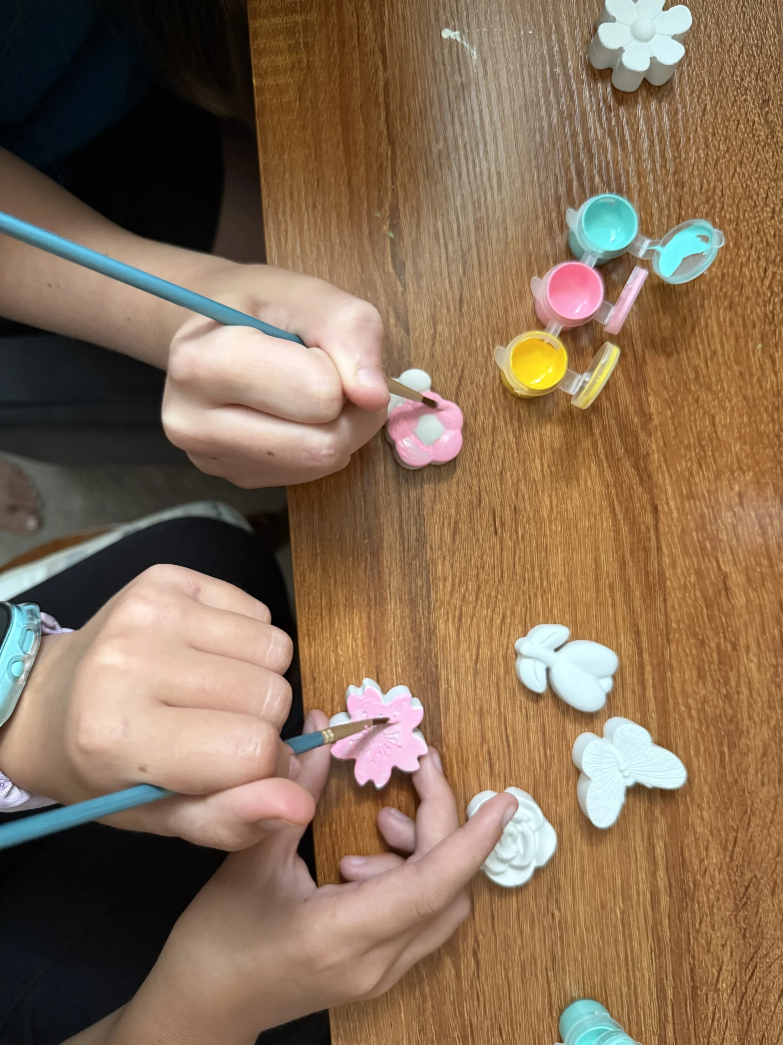 Children paint plaster butterfly and flower craft decorations at a wooden table.