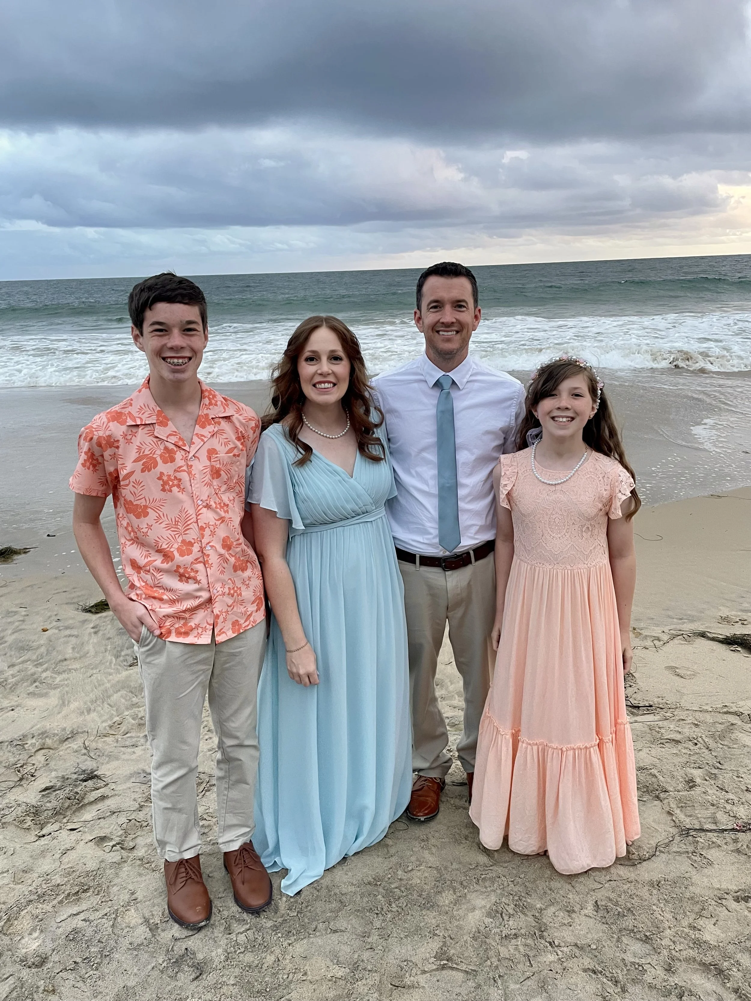 A family of four standing on a beach with ocean and cloudy sky in the background. The father, mother, son, and daughter are smiling and dressed in semi-formal summer clothing.