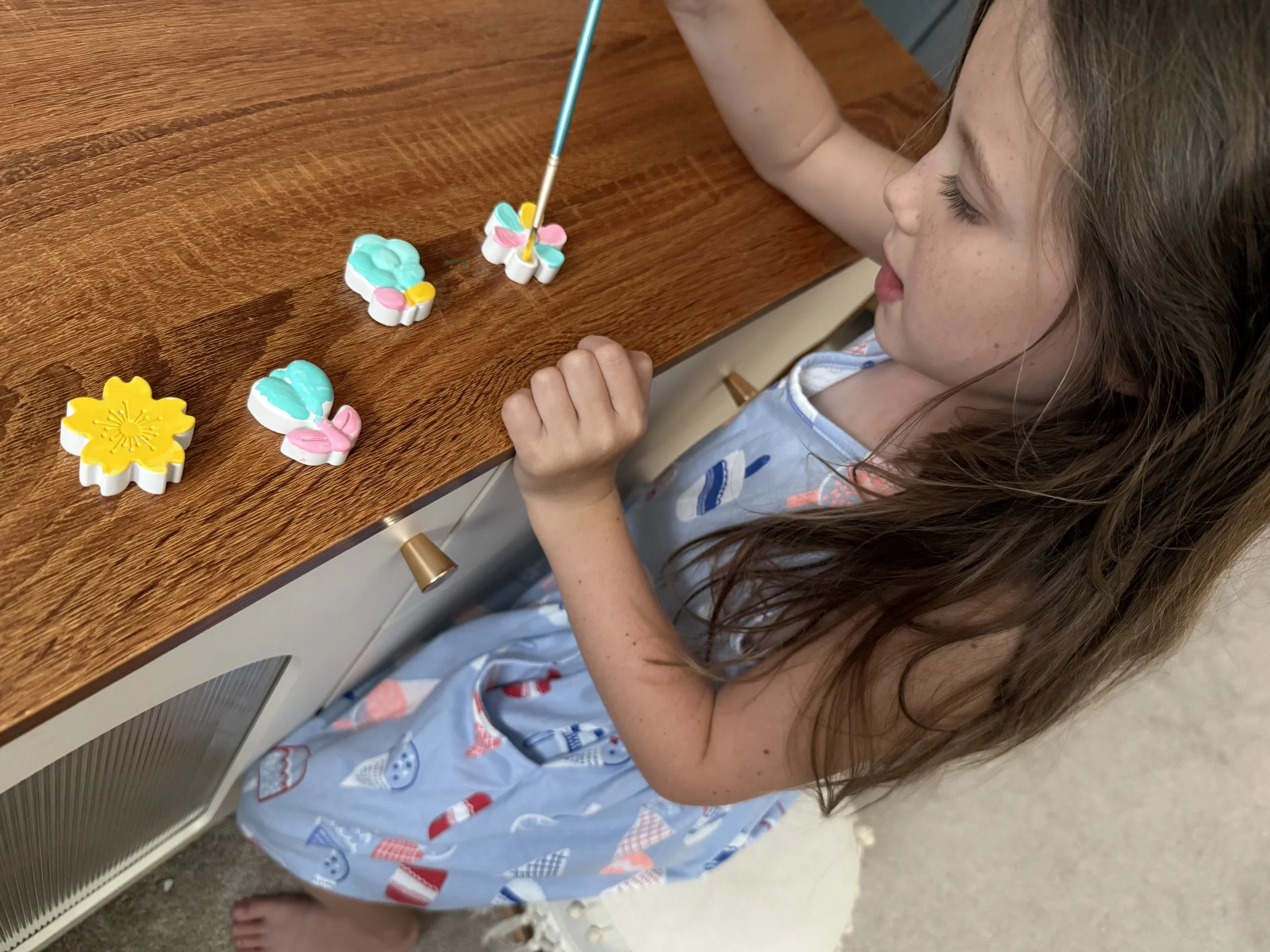 A young girl with long brown hair and freckles sits at a wooden table, painting bright-colored paint onto cute, flower-shaped plaster pieces with a wooden paintbrush.