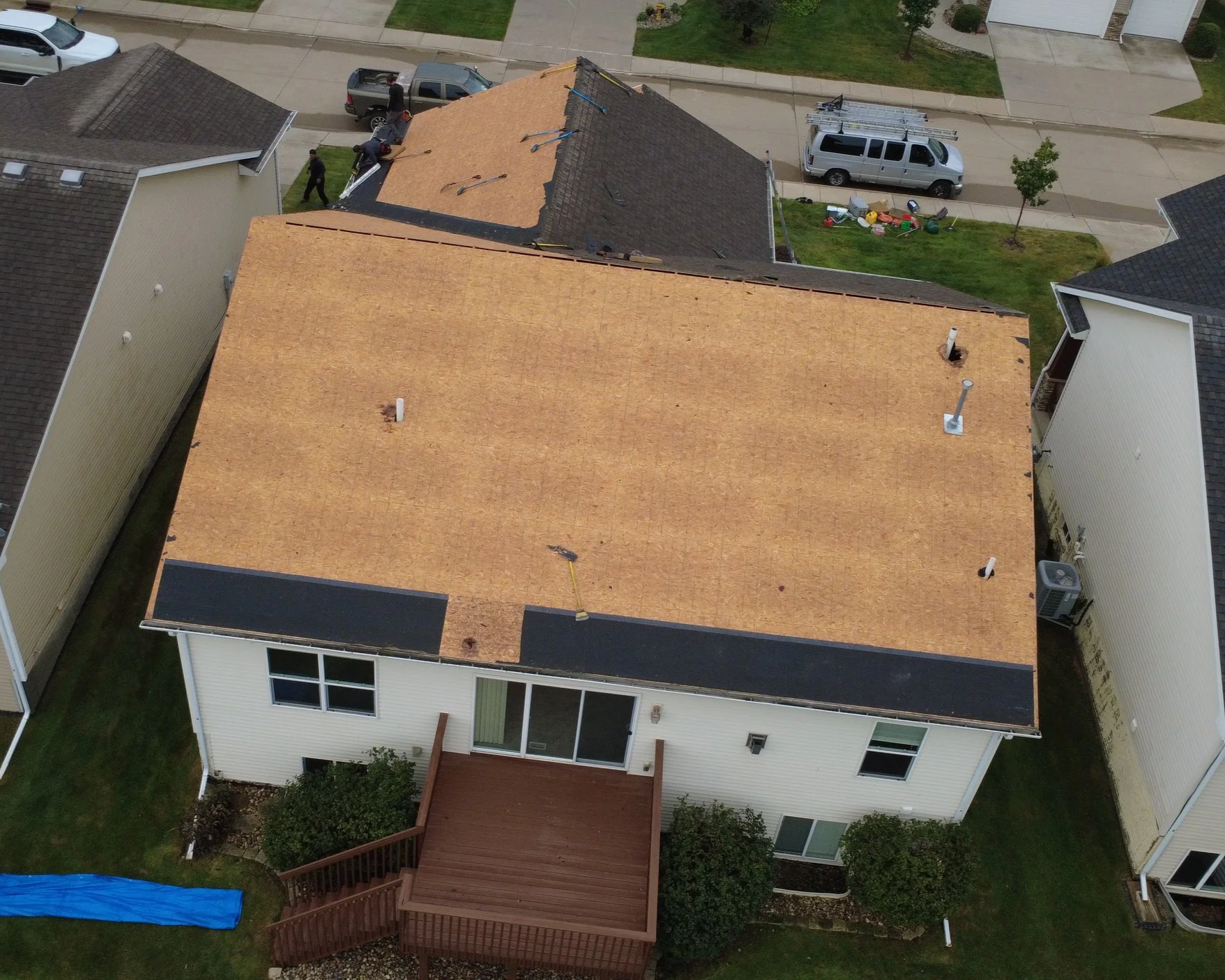Aerial view of a house with a new roof being installed, showing workers on the roof, trucks parked nearby, and neighbor houses with greenery and lawns.