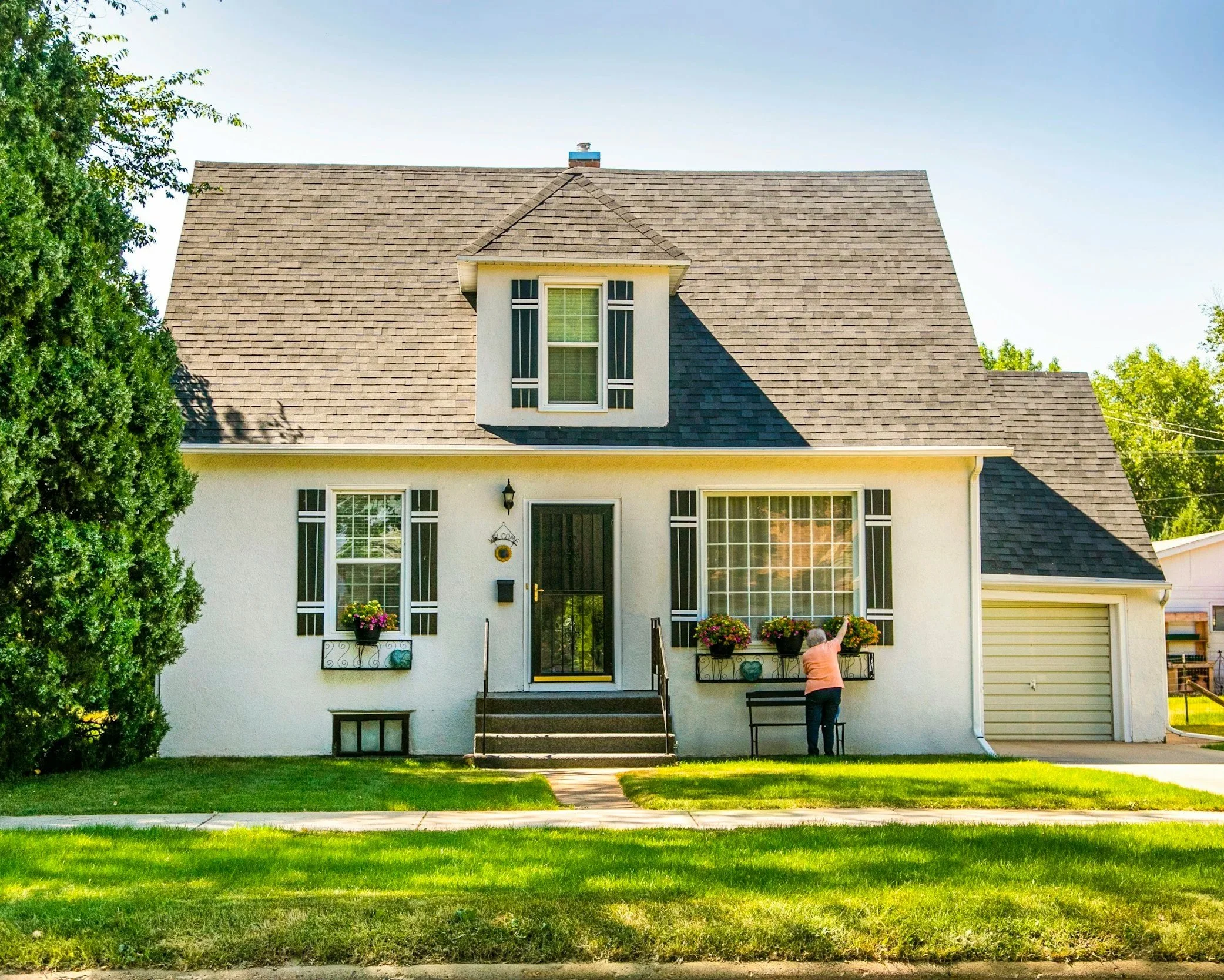 A person is standing on the sidewalk, reaching up towards a window of a two-story house with a pitched roof and dormer window, surrounded by green grass and trees.